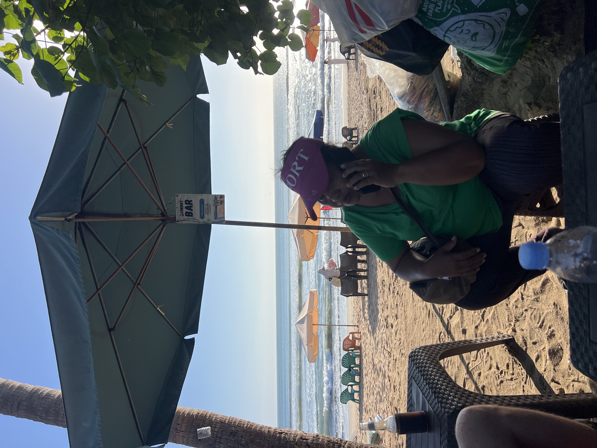 A woman in a green shirt and a purple visor is sitting on a beach in Kuta, Indonesia, talking on her phone. She is crouched on the sand under a large green umbrella, with a plastic bag and other items beside her. In the background, the ocean with waves is visible, along with several beach chairs and umbrellas. The sun is shining brightly, casting long shadows. A bottle of dark liquid and a bottle of water are on tables in the foreground.