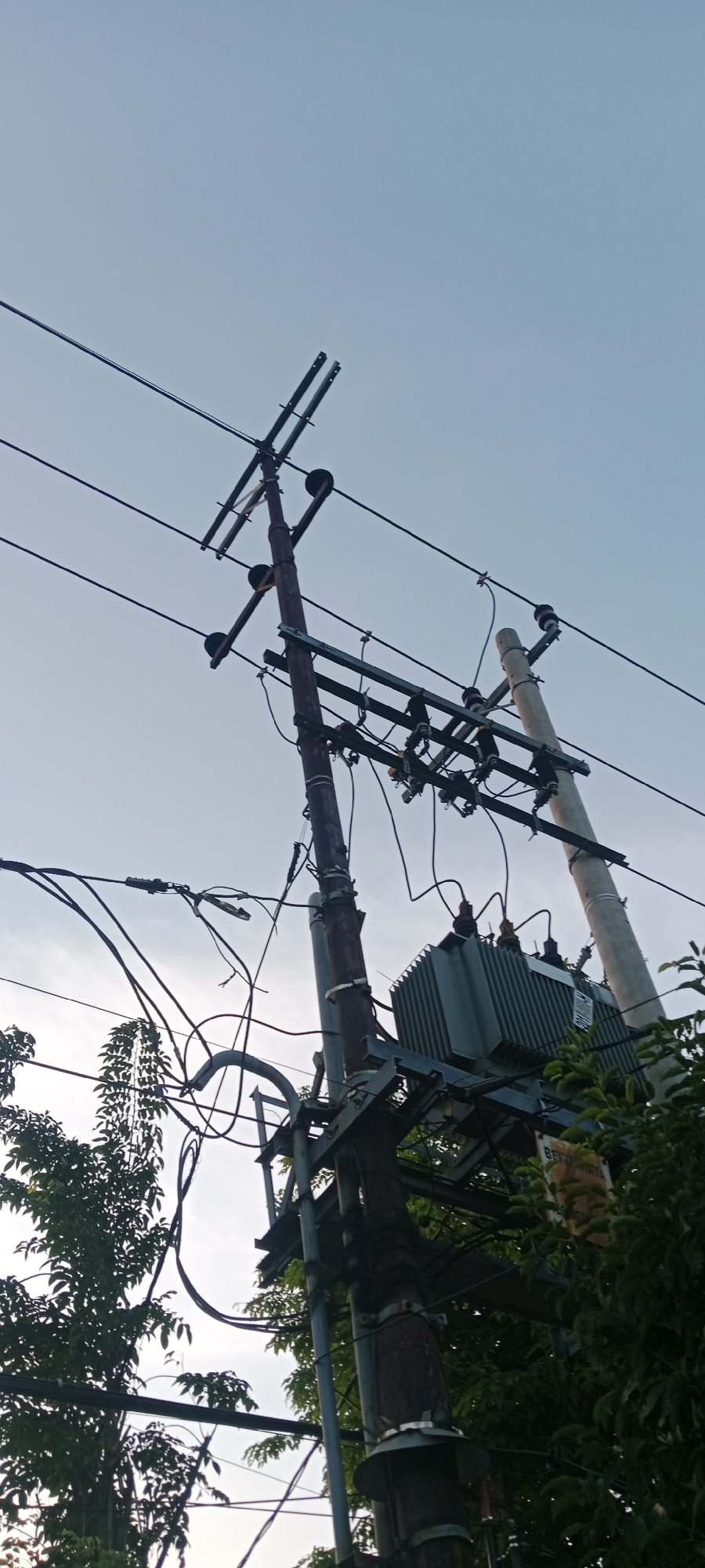 The image shows a utility pole with a transformer and electrical wires against a pale blue sky. The pole is made of dark metal and is positioned at an angle, with several crossbars supporting wires. A large, gray transformer box is mounted on a metal structure attached to the pole. Another, lighter colored pole is visible to the right, also supporting wires. The scene is partially obscured by lush green foliage from trees on the left and right sides of the frame. The photograph appears to be taken from a low angle, looking upwards. The location is identified as Tuban, Indonesia. There are no people or animals visible in the image.