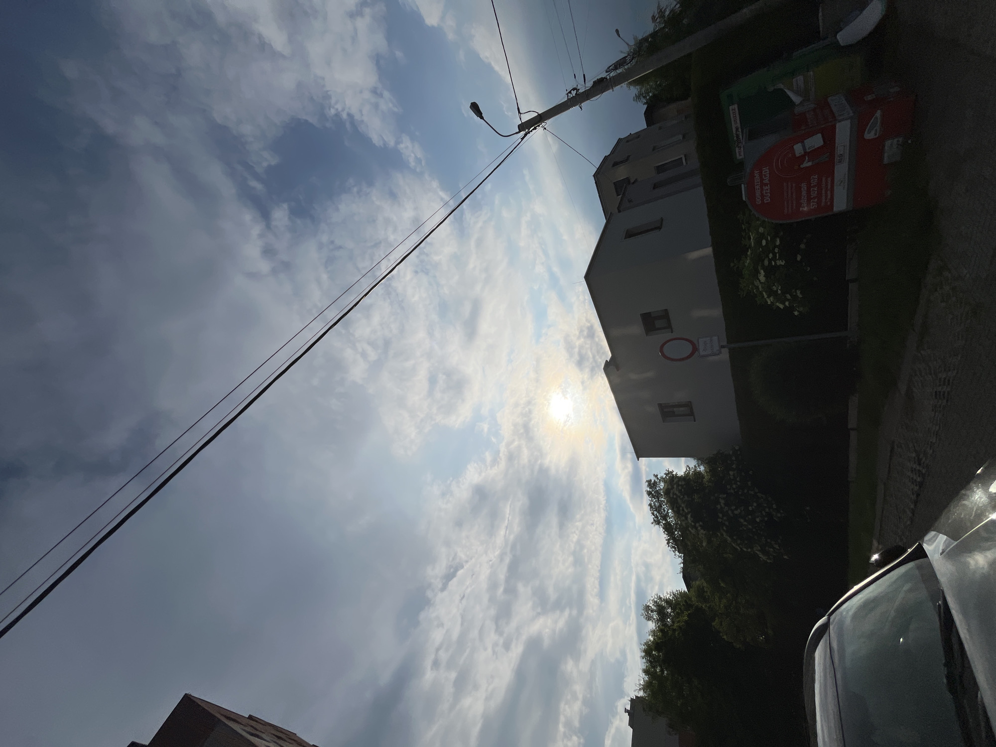The image, originally rotated 90 degrees clockwise, depicts an outdoor street scene looking upwards towards a bright, partly cloudy sky. The sun, partially obscured by white clouds, is a strong light source in the upper center, casting a bright glare. Patches of blue sky are visible between the clouds.
In the mid-ground, a tall utility pole with a streetlamp extends from the right, supporting multiple dark electrical wires that stretch across the frame towards the left. Behind the pole, a multi-story, light-colored building with dark windows stands. Below it, lush green foliage and trees are visible. Further to the right of the building, a large red collection bin or container is present, adorned with text. On the ground level near the building, a circular red traffic sign with a white horizontal bar (typically indicating 'No Entry' or similar restriction) is mounted on a pole, with a smaller white rectangular sign below it whose text is unreadable. The top of another building is visible on the far left edge of the frame.
In the lower right foreground, the hood and windshield of a dark-colored car are partially visible, reflecting the bright sky.
Visible text on the red collection container includes Polish words such as "GÓRZEŃCZYNY", "DUŻE AGD" (likely referring to large home appliances), and "Zadzwoń: 572 632 112" (Call: phone number).
The scene captures a bright daytime moment under a dynamic, cloudy sky. No specific activity or people are visible, presenting a static urban or suburban street view.