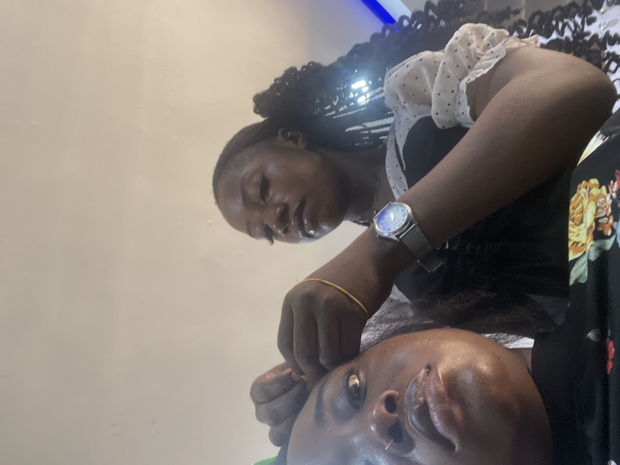 The scene depicts two individuals within an indoor environment in the city of Abuja, Nigeria. In the immediate foreground, the upper face of a person with dark skin is visible, featuring glossy lips and a silver nose ring on the left nostril. This individual wears a black garment with a floral pattern visible on the lower left. Partially obscured and positioned behind and to the right, a second person with dark skin is actively engaged in hair styling, with their hands positioned near the scalp of the foreground individual. This second person wears a black top with white, sheer, polka-dotted puffed sleeves, and has long, dark braided hair styled over their right shoulder. A silver-toned wristwatch with a circular face and a thin yellow bracelet are visible on their left wrist. The background is a plain, light-colored wall or ceiling, with a blue light source discernible on the far right.