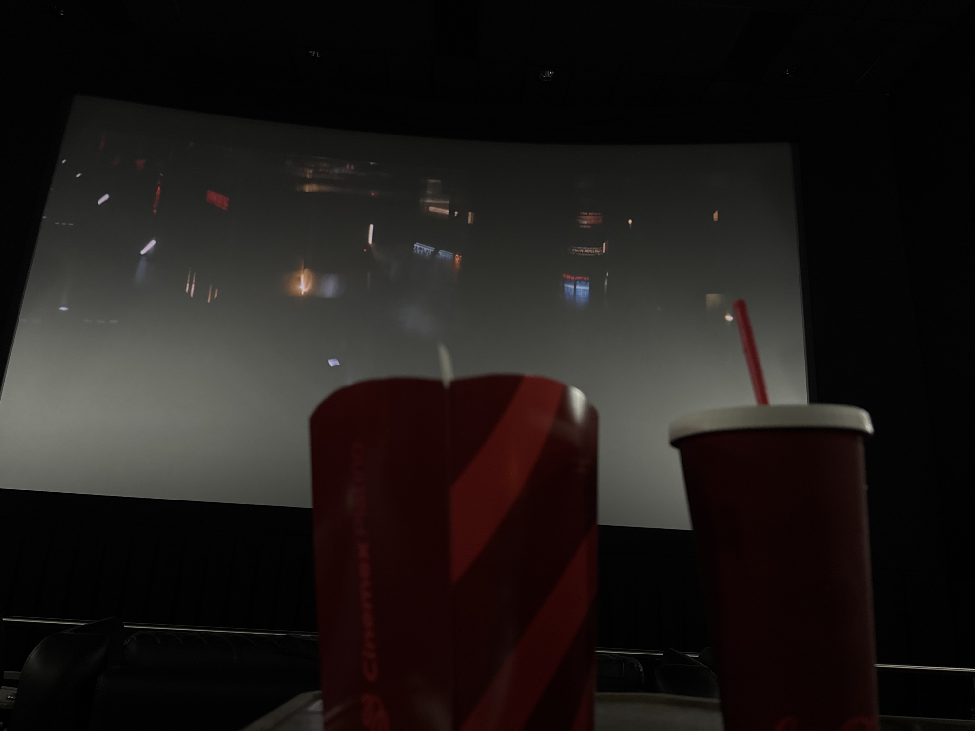 The photo depicts a scene inside a cinema in Mexico City, Mexico, featuring two red soda cups in the foreground. The cinema screen shows a blurry image of what appears to be an urban landscape at night. In the background, the cinema's seating is visible.