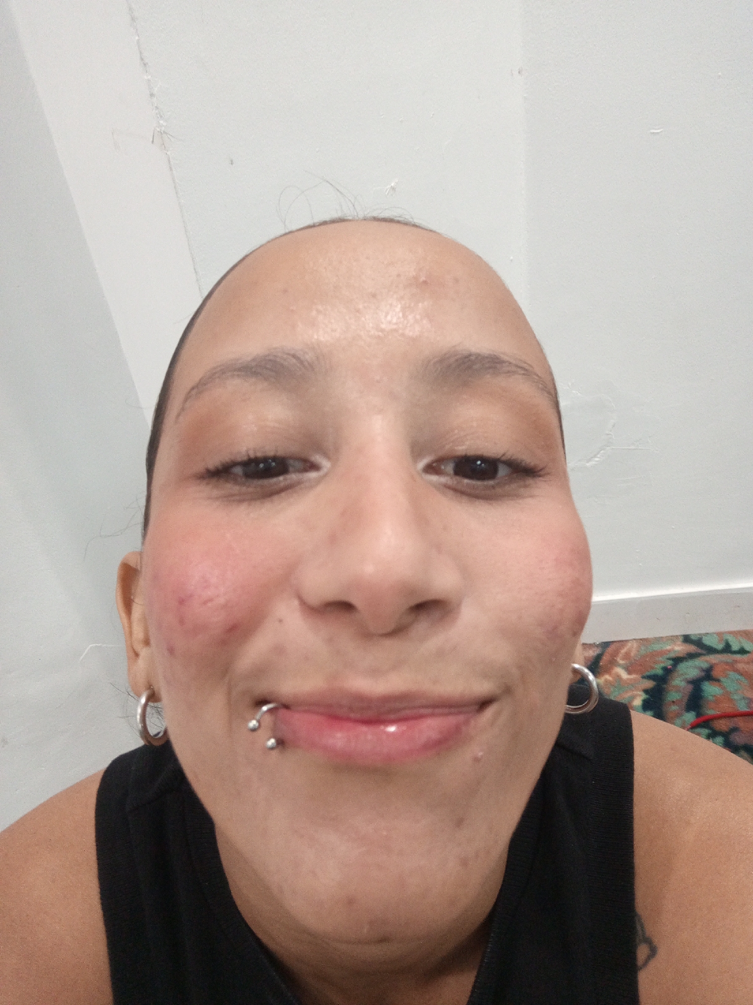 A close-up selfie captures a person with medium complexion, possibly of Hispanic descent, facing the camera directly. The person has dark eyes and eyebrows, a lip piercing with a horseshoe-shaped barbell on the left side of their lower lip, and hoop earrings. The background is a plain white wall, and a section of a patterned rug is visible at the bottom right corner. The person is wearing a black tank top. The scene appears to be indoors. There is no information provided that this is in Porlamar, Venezuela.