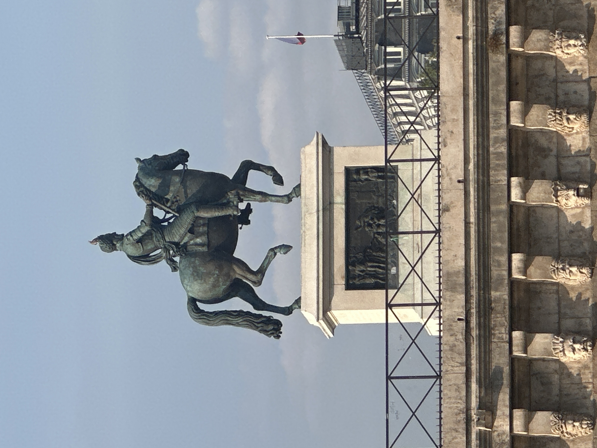 A full shot from below shows a bronze equestrian statue against a light blue sky with some clouds. The statue depicts a man in armor on a horse. The man is wearing a helmet with a plume and is holding reins. The horse is rearing up on its hind legs. The statue is on a tall, rectangular pedestal that has bas-relief carvings on its sides. In front of the pedestal is a black metal railing. Behind the statue and pedestal, a building with an ornate facade and a flagpole with a French flag can be seen. The bottom of the image shows a stone wall with decorative carvings of faces. The overall scene suggests an outdoor monument in a city.