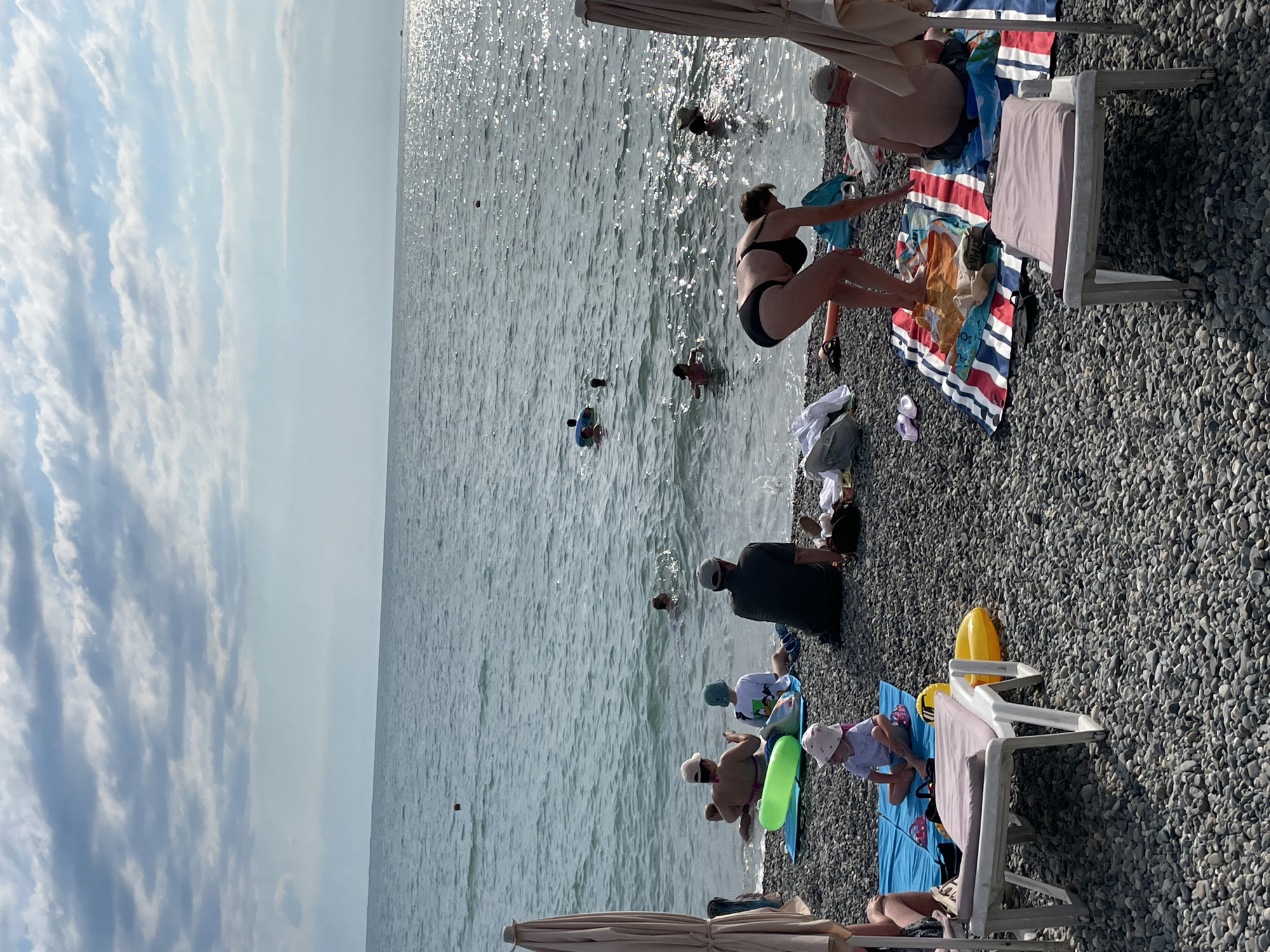 The image depicts a sunny day at a pebble beach in Sochi, Russia. Several people are visible, enjoying various activities. In the foreground, a woman in a black bikini is in the process of entering the water, with her arms outstretched. To her right, a man in a light-colored cap and shorts sits on a striped towel, leaning back on his hands. Further down the beach, several children and adults are either playing in the shallow water or sitting on towels. One child is wearing a white t-shirt and a blue hat, while another is in a striped shirt and a white hat. A yellow inflatable toy is also visible. In the background, the sea stretches out, with sunlight glinting off the water. The sky is partly cloudy, with soft white clouds against a pale blue backdrop. The overall atmosphere appears relaxed and leisurely, suggesting a typical summer day at the beach.