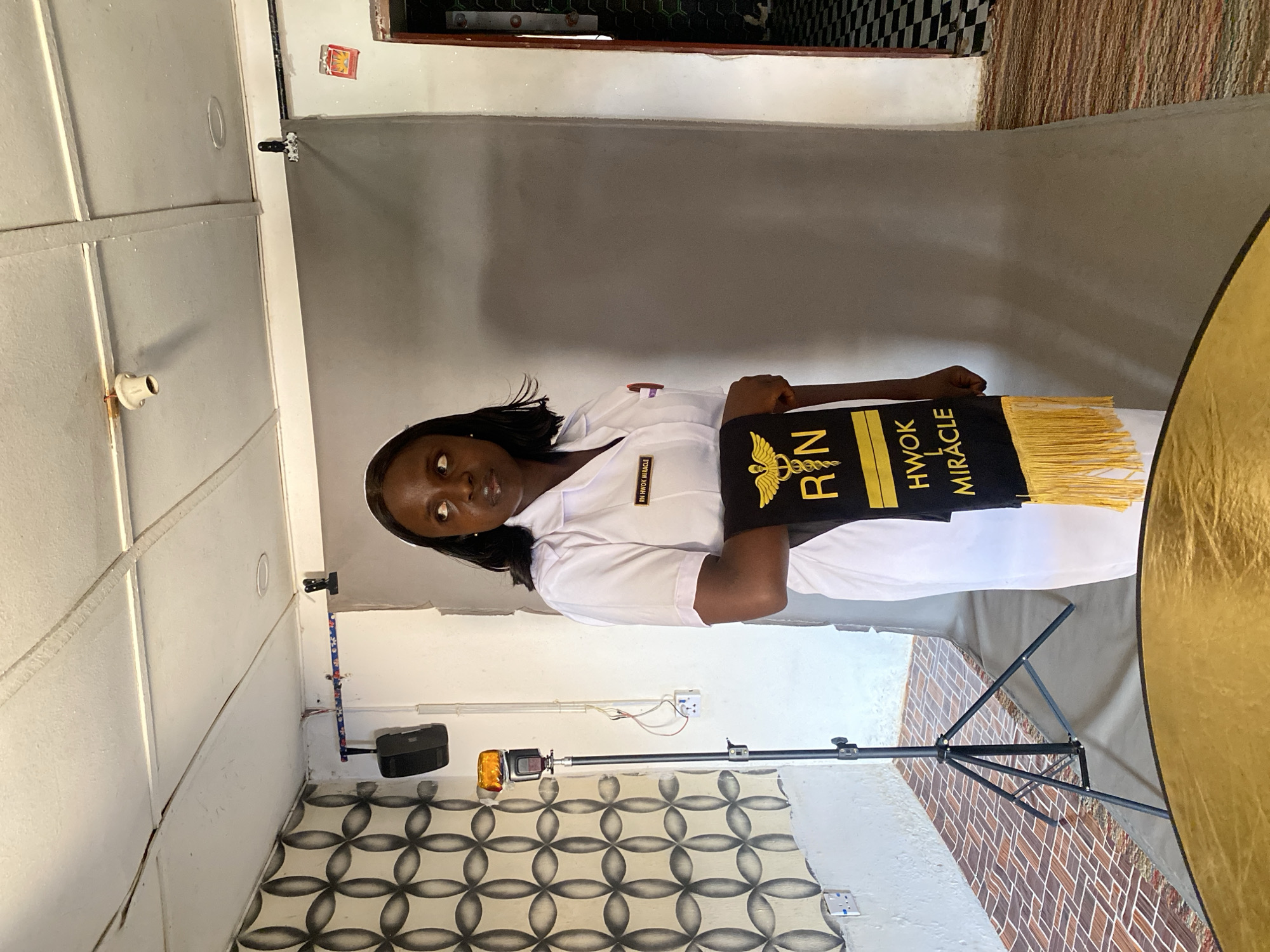 A young woman in a white nurse's uniform stands in a room holding a black and gold graduation sash with the letters "RN" and the name "HWOK MIRACLE". The sash has gold tassels. The woman is facing the camera. The room appears to be a makeshift studio with a gray backdrop and lighting equipment. The floor has a patterned tile design. The location is likely a building in Vom, Nigeria.