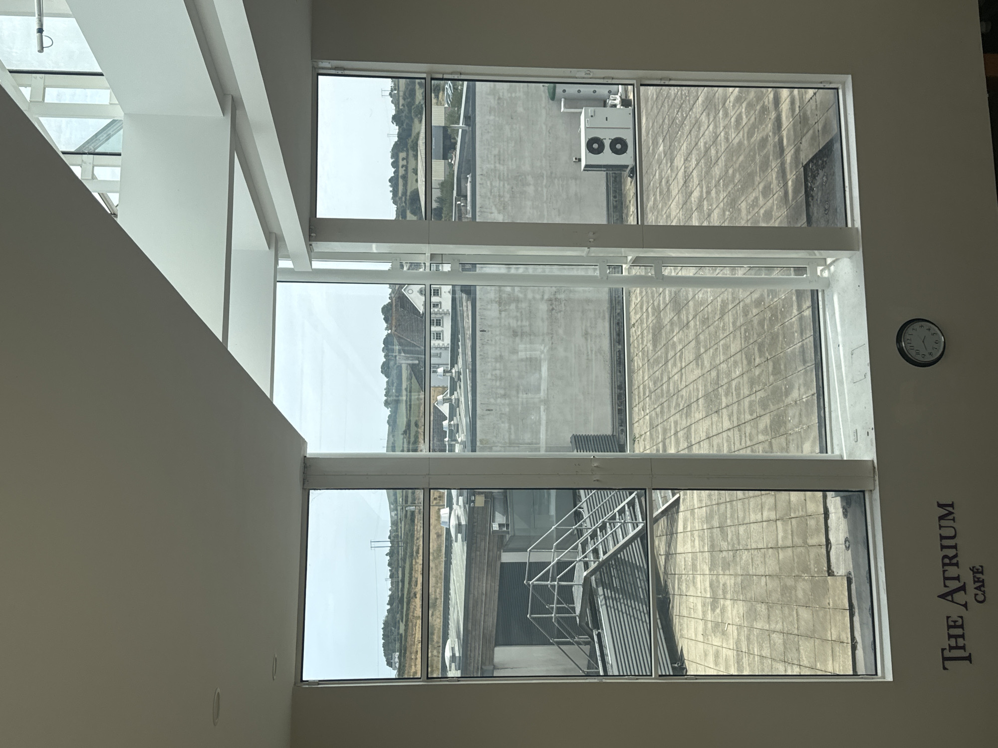 The image shows a view through a large window of an interior space overlooking an outdoor industrial area and a distant rural landscape. The interior space has a white ceiling with recessed lighting. Below the window, text on the wall reads "The Atrium Café". A round clock is mounted on the wall to the left of the window.
The outdoor view through the window includes a flat, paved area with square tiles, interspersed with some darker, wet-looking patches. A metal staircase with railings leads down to a corrugated metal structure. In the background, there are several industrial buildings with flat roofs, some with ventilation units or chimneys. Further in the distance, rolling green hills and fields are visible under a cloudy sky, with some scattered houses and telephone poles. The overall impression is of a modern building with a view of its surrounding environment. The location is identified as Unknown, Ireland.