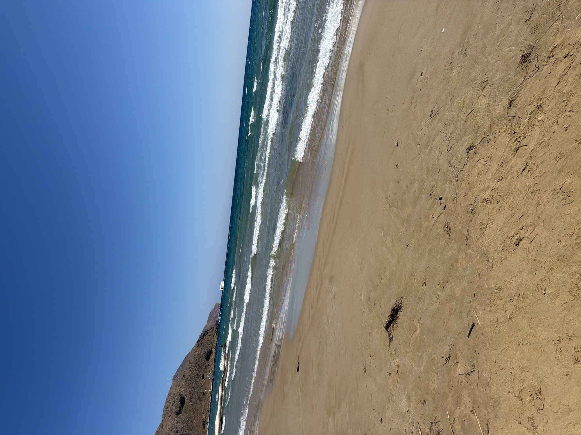 A wide-angle view of a sandy beach in Georgioupoli, Greece, under a clear blue sky. The foreground is dominated by light brown sand with scattered debris, including dried seaweed and small sticks. In the middle ground, gentle waves with white crests break and recede onto the shore. The sea is a deep turquoise color. In the background, a chain of rocky, brown hills slopes down towards the water. A white structure, possibly a building or a boat, is visible on the far right of the water. A few indistinct figures are present in the shallow water near the shore. The overall scene depicts a serene coastal landscape on a sunny day.
