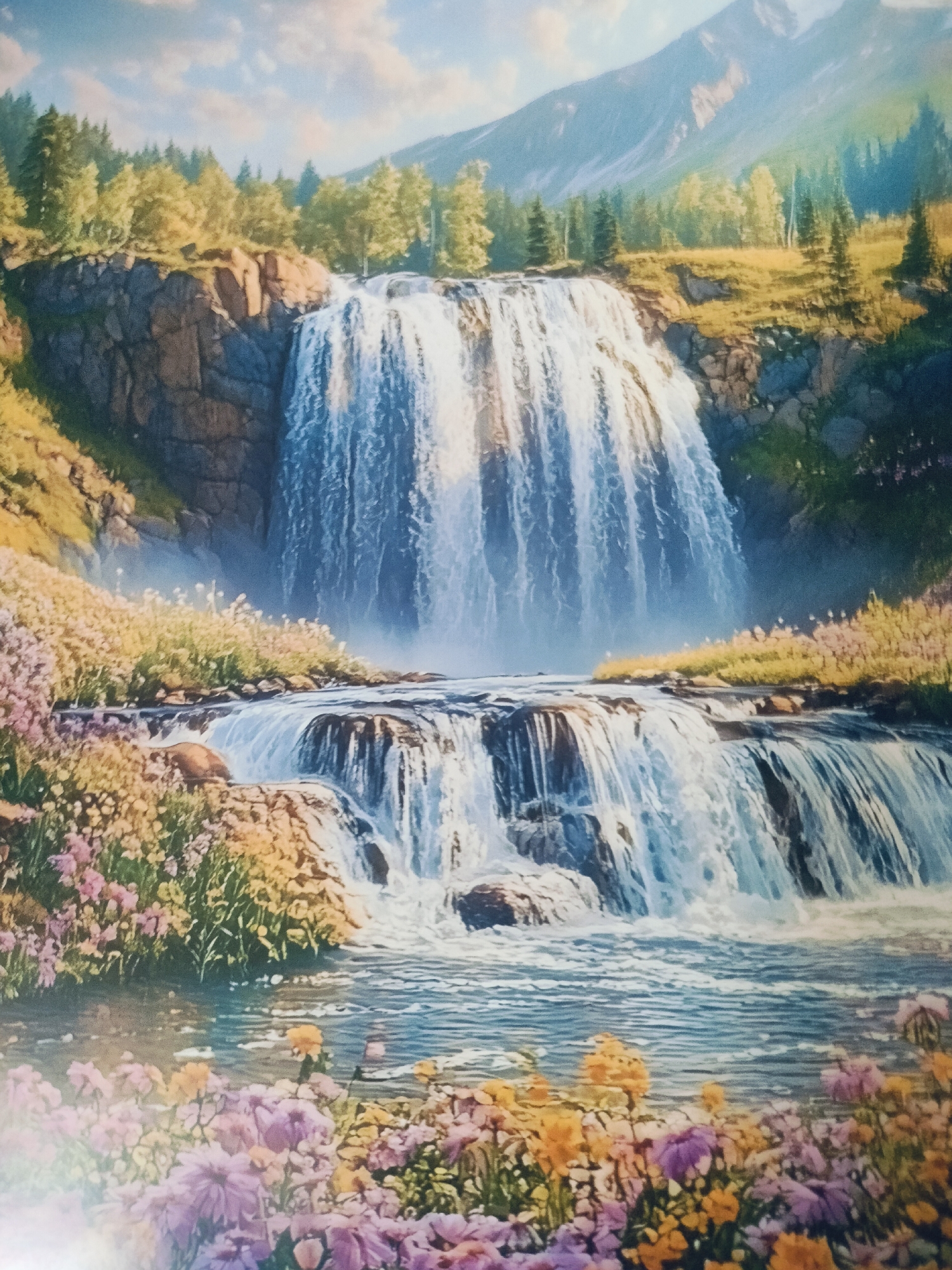 The image depicts a picturesque natural landscape featuring a multi-tiered waterfall cascading down rocky cliffs. Lush green trees and vegetation surround the upper portion of the falls, while a serene body of water collects at its base. The foreground is adorned with a vibrant array of wildflowers in various shades of pink, purple, and yellow, adding a splash of color to the scene. In the background, majestic mountains rise, their peaks partially obscured by soft clouds. The sky above is a clear blue, with a few wispy clouds scattered across it, suggesting a pleasant day. There are no people or man-made structures visible in the image, emphasizing the untouched beauty of the natural environment. The overall impression is one of tranquility and natural splendor. This specific scene is not identifiable as being located in the city of Aleysk, Russia.