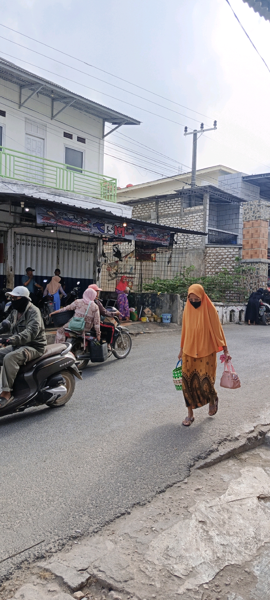 An outdoor street scene in Gayam, Indonesia, depicts a woman wearing an orange hijab and patterned skirt walking across the asphalt road, carrying a pink handbag in her right hand and a green and white striped bag in her left. She is walking from right to left in the frame. In the background, several people are seen on or around motorcycles. A man in a grey jacket and hat is riding a motorcycle on the left side of the road. Behind him, two women on a motorcycle are also present. Shops with corrugated metal shutters and banners line the left side of the street, with some merchandise visible outside. Buildings with brick and concrete facades are visible in the background. Power lines stretch across the sky, and a utility pole is present. The road surface appears somewhat uneven and worn.