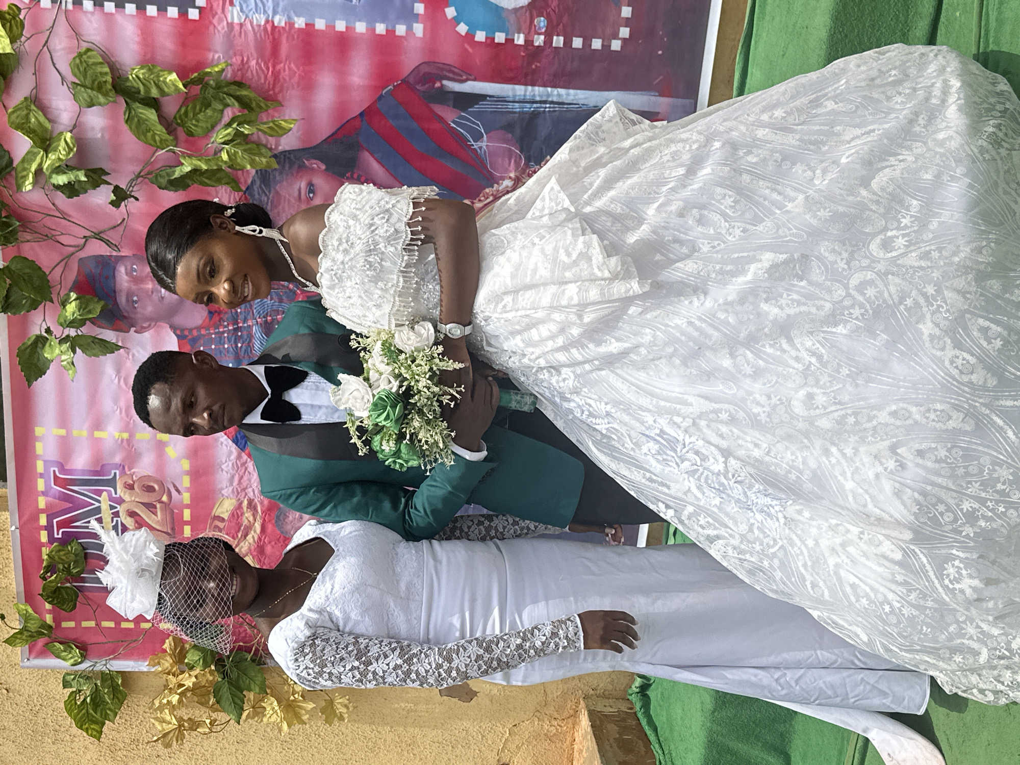 The image depicts a wedding portrait. A man in a green suit and bow tie stands next to a woman in a white wedding gown, holding a bouquet of white and green flowers. Behind them is a backdrop featuring an image of another woman in a white dress and a man in casual wear. The backdrop also has decorative foliage and the number "28" on it. The scene appears to be set in an unspecified location in the city of Unknown, Unknown. The subjects are posing for a photograph.