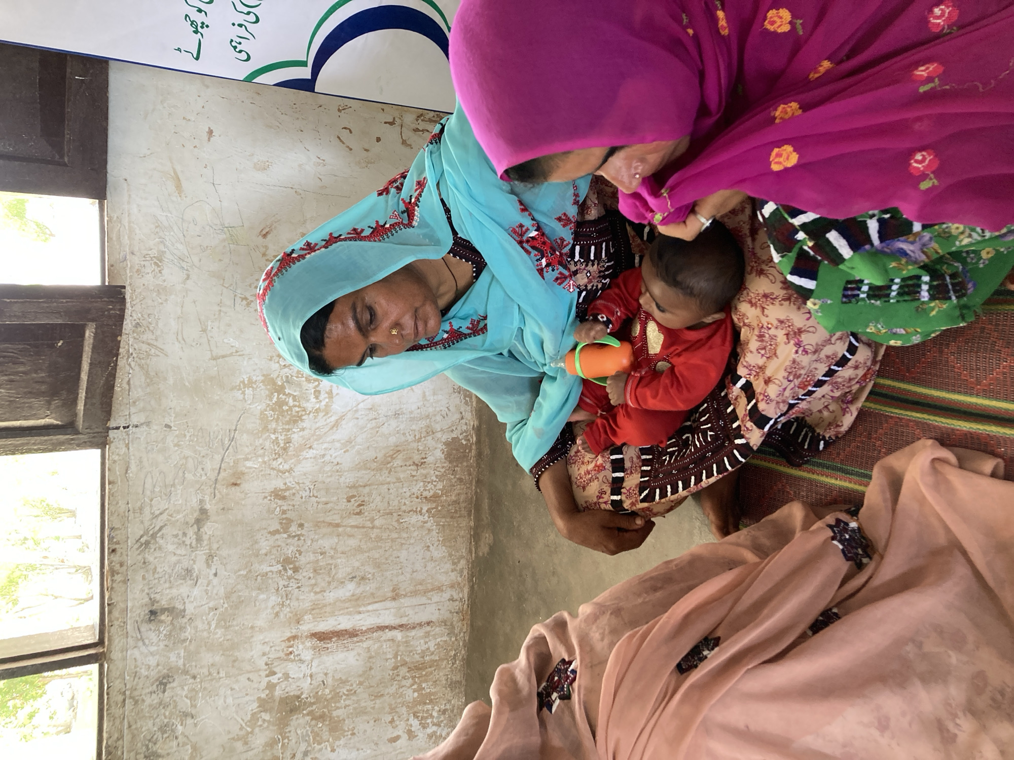In Goth Ali Marād, Pakistan, a woman in a blue embroidered headscarf and brown dress holds a baby in a red shirt. The baby holds a green and orange bottle. A second woman in a purple headscarf is close to the baby. The women are seated on a mat, and a peach-colored cloth is nearby. A window is visible, and the wall is worn.