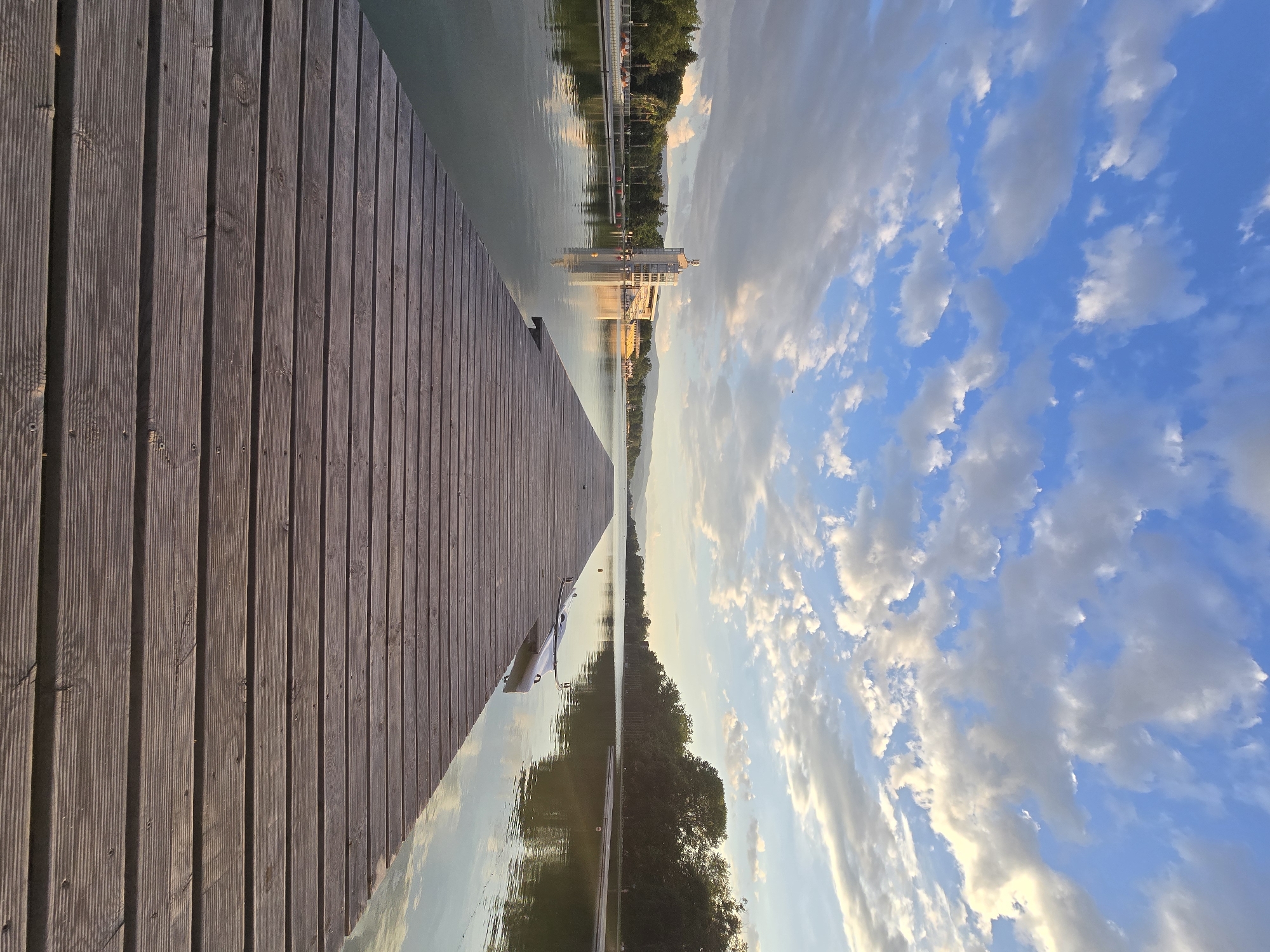 The image shows a dock extending into a body of water. The dock is made of wooden planks, with the planks running parallel to the direction of the dock's extension. To the right of the dock is water, which reflects the sky above. The sky is blue with scattered white clouds. In the distance, there are trees and what appears to be a building on the water. The image is taken from a low angle, looking out over the water. Location and city are unknown.