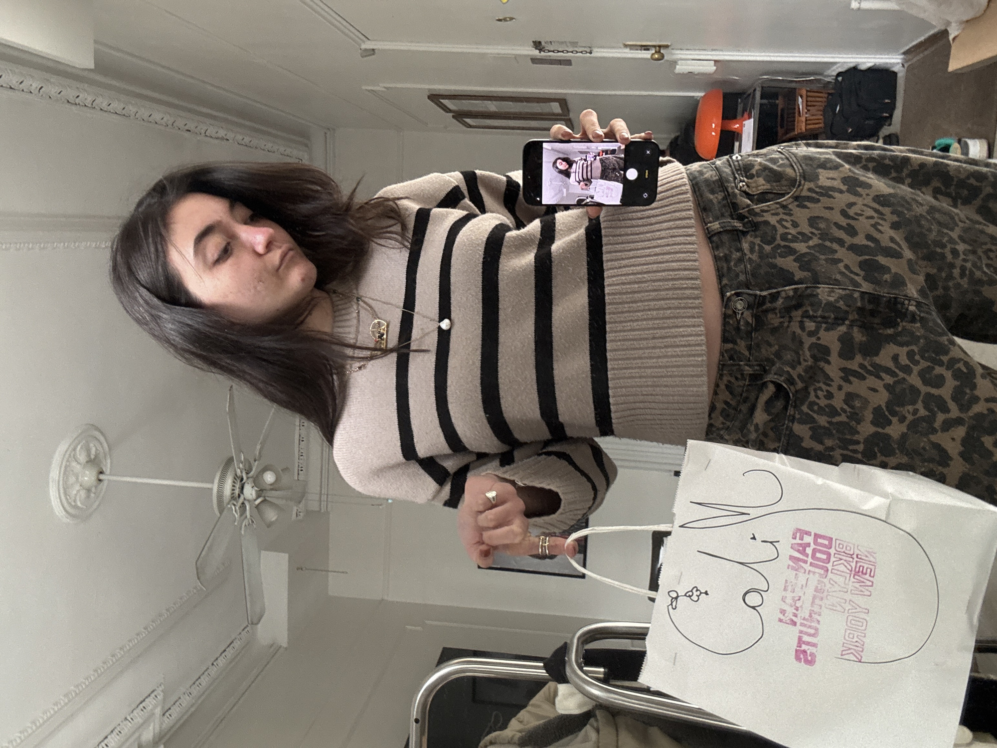 An indoor mirror selfie, captured with a 90-degree clockwise rotation, shows a young woman in profile. She has long, dark hair and a serene expression, with her eyes cast downwards. She is wearing a cream-colored sweater with broad black horizontal stripes and dark leopard print pants.
The woman holds an iPhone in her right hand, positioned to photograph her reflection. The phone screen clearly displays the same scene, showing her attire and posture. Her left hand holds a white paper bag. She wears a long necklace with a small, circular pendant and several rings on her fingers, including a heart-shaped ring and another with a double band.
The background reveals a room with a white ceiling adorned with decorative molding and a white ceiling fan with multiple blades. Other elements visible in the background include a doorknob, and various indistinct household items or storage solutions.
The white paper bag held by the woman features a hand-drawn illustration resembling a stylized face with a prominent nose and lips, along with a small flower drawing. Visible, reversed text on the bag reads "FAMILY BAKER & DONUTS" and "NEW YORK", indicating the bag's origin from a New York establishment. The overall scene captures a candid, everyday moment indoors.