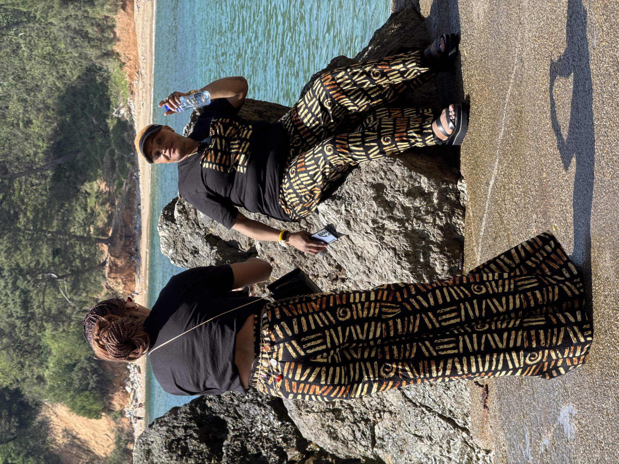 Two people wearing matching black tops and patterned trousers are sitting on rocks near a beach in Budva, Montenegro. The person on the top is reclining with a bottle of water in their hand and holding a cell phone. The other person is sitting with their back to the camera and has a purse. The background shows a view of the Adriatic Sea, a sandy beach, and green trees on a rocky cliff. The lighting suggests it is a sunny day.