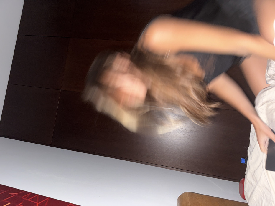 A blurry image shows a woman with long brown hair and a black shirt. She's lying on a bed with white sheets and holding a black phone or tablet. A dark brown wooden headboard is behind her. The background also shows a white wall and part of a red patterned object on the floor. The image is poorly focused, making details difficult to discern.