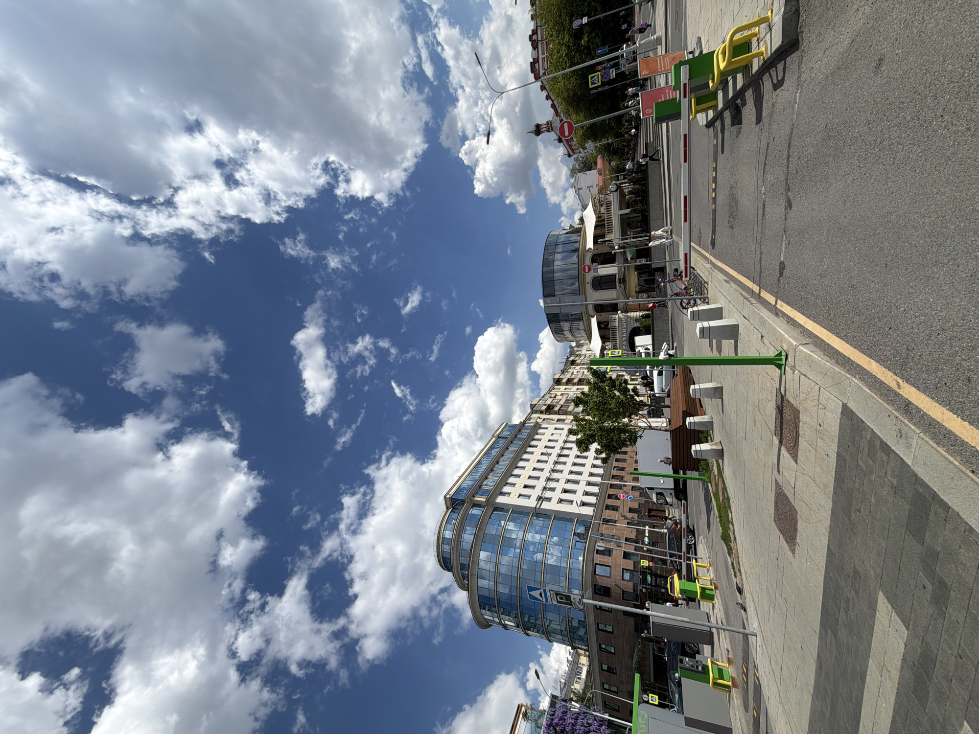 This daytime image captures a vibrant urban street scene, likely in Moscow, Russia, under a bright blue sky filled with scattered white cumulus clouds. The sun is shining, casting clear shadows on the ground.
In the foreground, a paved street with a yellow dividing line runs parallel to a wide sidewalk made of patterned grey and darker grey paving stones. Green and yellow metal barriers and bollards are strategically placed along the street and sidewalk edges. Street infrastructure includes lampposts, traffic signs, and a red and white boom barrier.
On the left, a prominent modern multi-story building with a distinctive curved glass facade reflects the sky and clouds. Its upper floors are light-colored. A tree with green foliage stands next to this building, and purple flowering plants are visible in the lower left. The street is lined with various buildings, showcasing a mix of architectural styles, including older structures. In the background on the right, a building with a distinct domed or pointed roof structure, possibly a church or a decorative element, adds a historical touch to the urban landscape.
A few vehicles, including a white van and a dark car, are partially visible on or near the street. Several indistinct figures of people can be seen walking on the sidewalk in the background, engaging in ordinary daily activities.
Visible Cyrillic text includes "НЕПРАВДИВЫЙ" (untruthful/false) on a green sign and "ДЕТИ" (children) on another sign further down the street, providing strong location cues consistent with a Russian-speaking city. The scene presents a bustling yet orderly urban environment.