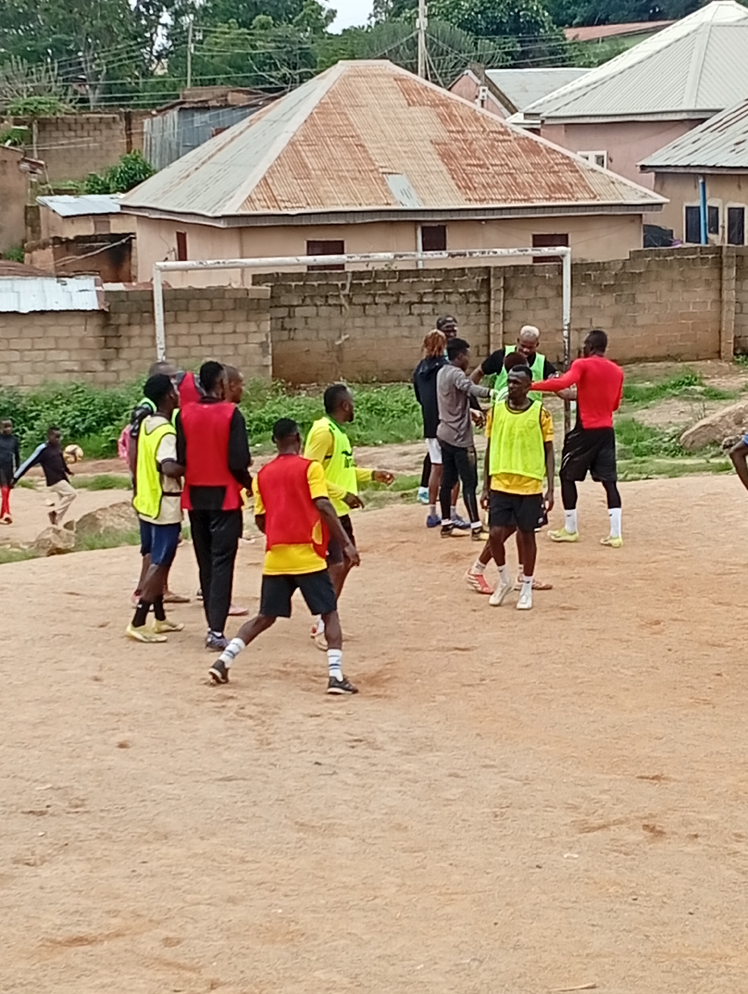 The image depicts an outdoor scene in Jos, Nigeria, where a group of young men are engaged in what appears to be a soccer practice. The ground is a dusty, dirt field, typical of many urban areas in Nigeria. In the background, residential buildings with corrugated iron roofs are visible, suggesting a mixed urban and semi-rural environment.
Several individuals are clad in athletic wear. Some are wearing bright yellow and red vests, possibly indicating team affiliations or practice drills. One person in the foreground is wearing a yellow vest over a yellow t-shirt and black shorts, with white socks and athletic shoes. Another individual in the mid-ground is wearing a red vest over a black shirt and black pants, also with athletic shoes.
A soccer ball is present in the scene, with one person appearing to kick it. The overall atmosphere suggests an informal sporting activity. The presence of the buildings and the attire of the individuals are consistent with the environment of Jos, Nigeria, a city known for its plateaus and a blend of urban development and open spaces.