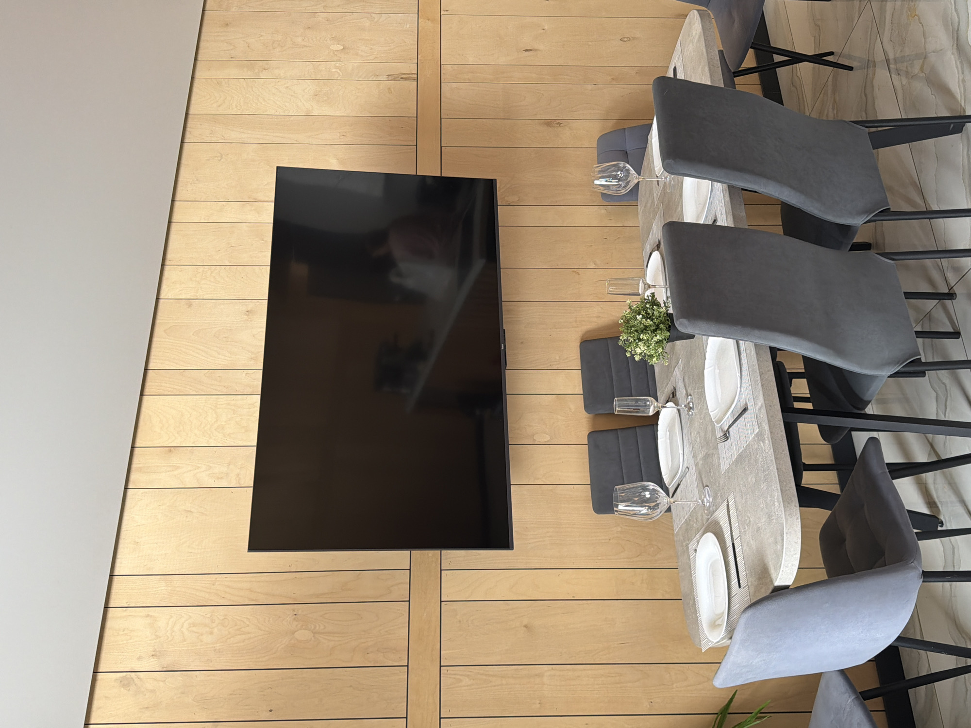 A dining area features a large, flat-screen television mounted on a light-wood paneled wall. In front of the wall, a modern dining table is set with white plates, cutlery, wine glasses, and a small potted plant. Several dark gray upholstered chairs with black legs surround the table. The flooring appears to be polished tile. No people are clearly visible in the scene, though a faint reflection of individuals can be seen on the television screen. The setting suggests an indoor dining space, possibly a restaurant or home, located in the city of Семилуки, Russia.