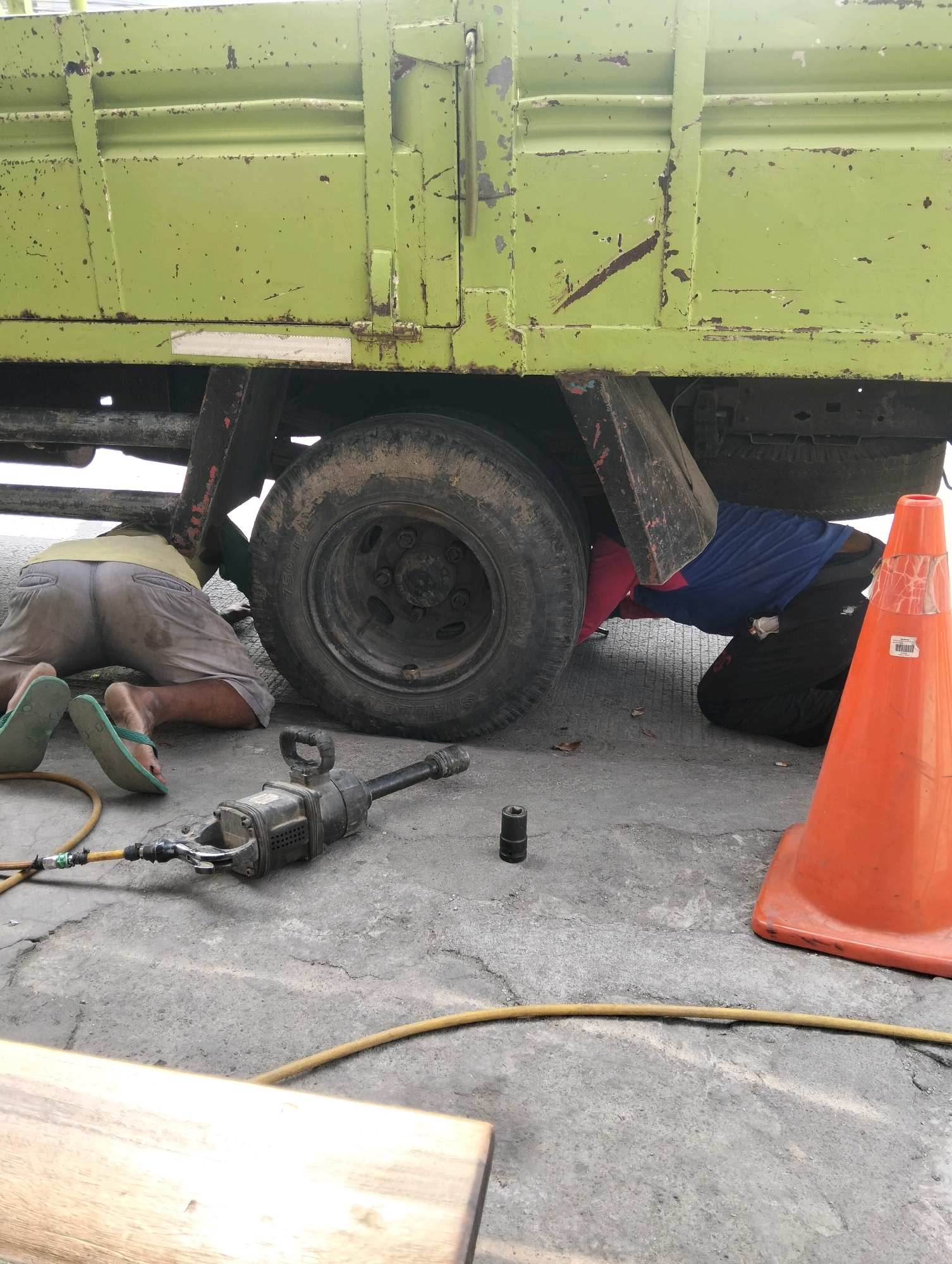In Semarang, Indonesia, two individuals are working on the underside of a green truck. One person, wearing light grey shorts and a yellow shirt, is on their hands and knees on the right side of the truck, facing away from the camera. Another person, wearing black pants and a blue shirt, is also on their knees on the left side of the truck, their body mostly obscured by the truck's undercarriage. An air-powered impact wrench and a yellow hose are visible on the ground near the first individual. An orange traffic cone stands to the right of the truck, and a piece of wood is in the lower left foreground. The ground is paved with concrete.