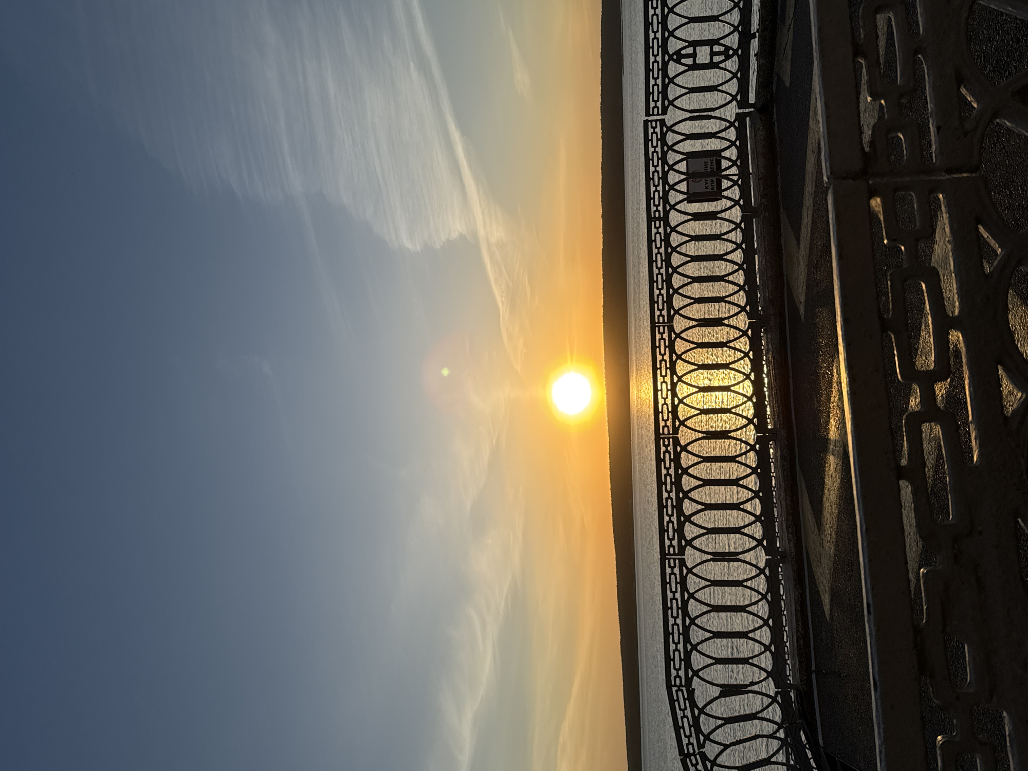 The image captures a sunset scene in Naberezhnye Chelny, Russia, viewed through an ornate metal fence. The sun, a bright yellow-orange orb, is positioned near the horizon, casting a golden glow across the sky and water. Streaks of white clouds are visible in the blue sky. The fence, constructed with a repetitive oval and geometric pattern, is partially silhouetted against the setting sun. The photograph is taken from a slightly elevated perspective, likely along a waterfront promenade or a bridge. The light conditions suggest the image was captured during the golden hour.