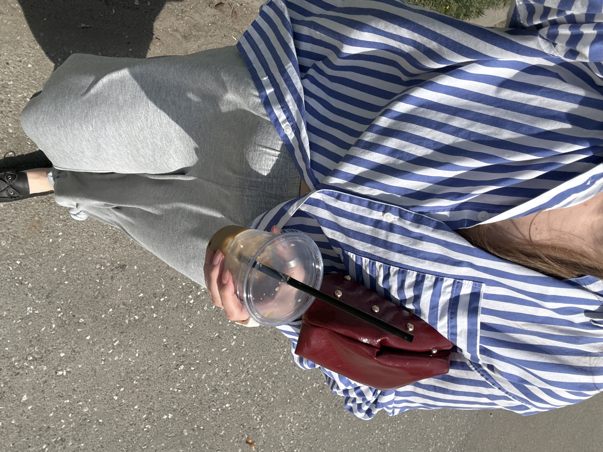 A woman stands on a paved surface, possibly a street, wearing gray sweatpants and a blue and white striped shirt. She holds a plastic cup with a black straw in her left hand. Her right hand rests on a small, maroon purse with metal studs. She is wearing black shoes. The location is not possible to identify as the location is not contained within the image.