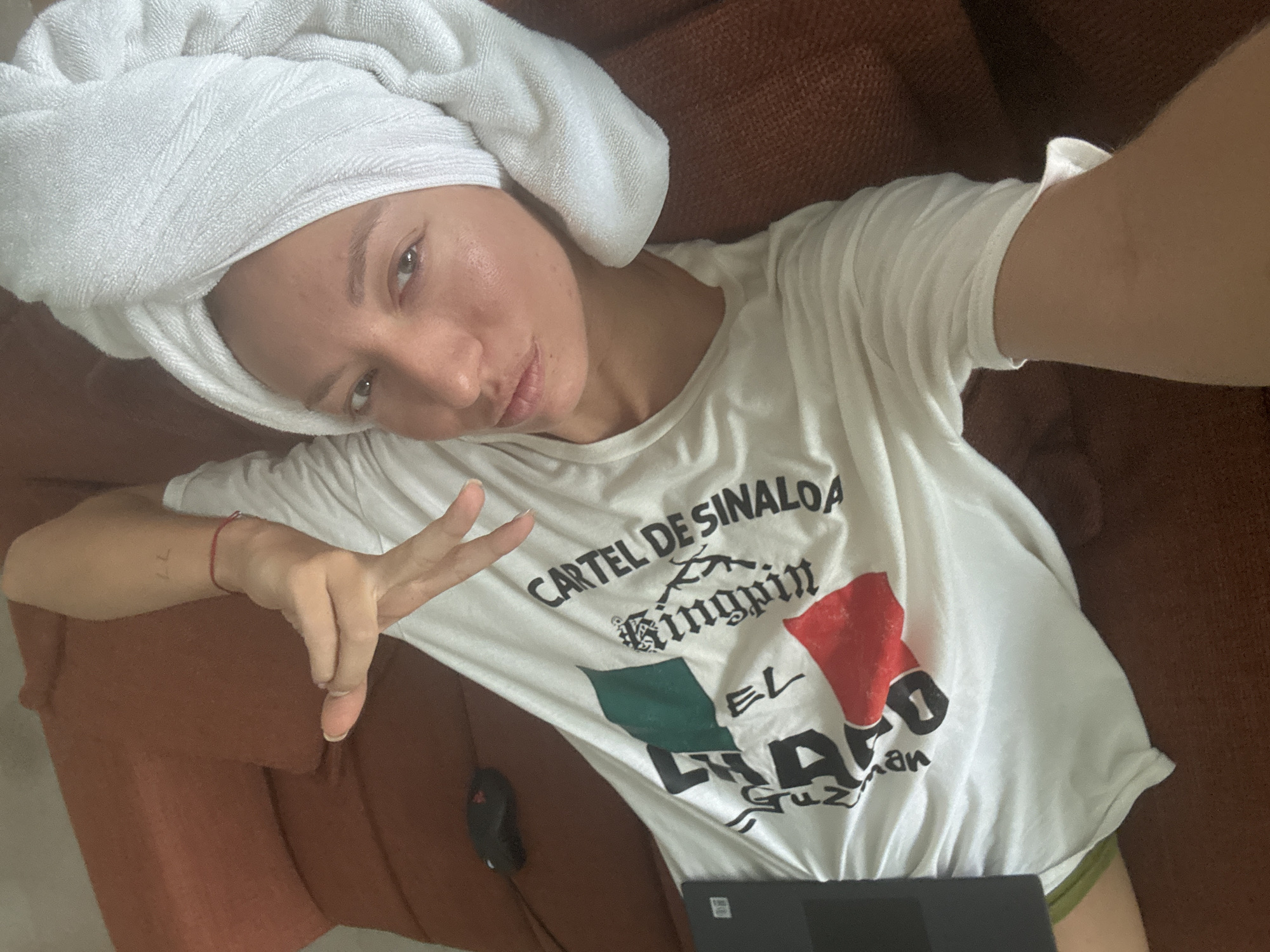 A woman wearing a white t-shirt with the text "CARTEL DE SINALOA" and a Mexican flag design is lying on a brown sofa. Her hair is wrapped in a white towel, and she is looking at the camera with her hand raised in a peace sign. A laptop and a computer mouse are on the sofa next to her. The background is an indoor setting with a textured brown sofa and wall. The image appears to be a selfie taken indoors. There is no information in the image to suggest it is located in Ko Samui District, Thailand.