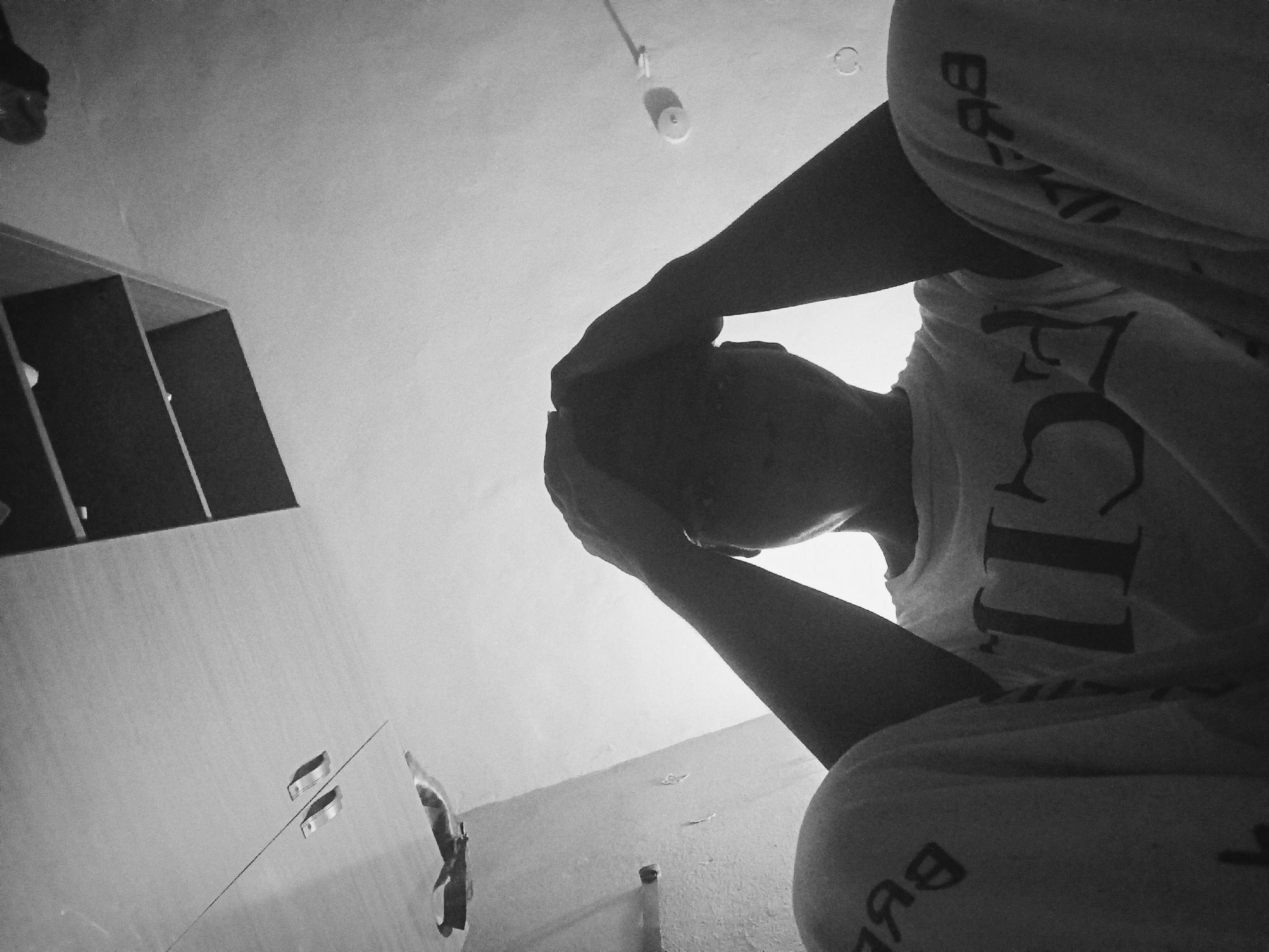 The monochrome image, captured from a low-angle, upward-looking perspective, depicts a solitary person indoors in what appears to be Nkpor, Nigeria. The individual is largely silhouetted against a bright background, with their head bowed and hands clasped over or resting on their head and face, suggesting a moment of deep introspection, sadness, or distress. They are wearing a light-colored t-shirt with dark text, where the letters "CII" are clearly visible on the chest. Their lower garment, possibly shorts or pants, is also light-colored and features mirrored dark text, including characters resembling "BB".
The setting is a room with minimalist furnishings. To the immediate left, a dark, multi-shelved unit, possibly a bookshelf or wardrobe, is partially visible. On the right, a plain, light-colored wall features what appears to be a cabinet or door with two distinctive silver-colored handles. The ceiling, prominent in the upper part of the frame, holds a small, round light fixture or bulb housing, and another circular attachment. The strong backlighting creates a stark contrast, emphasizing the person's silhouette and the emotional weight of their posture. The time of day and external weather conditions cannot be determined from the indoor, black and white photograph. The image conveys a profound sense of personal solitude and contemplation.