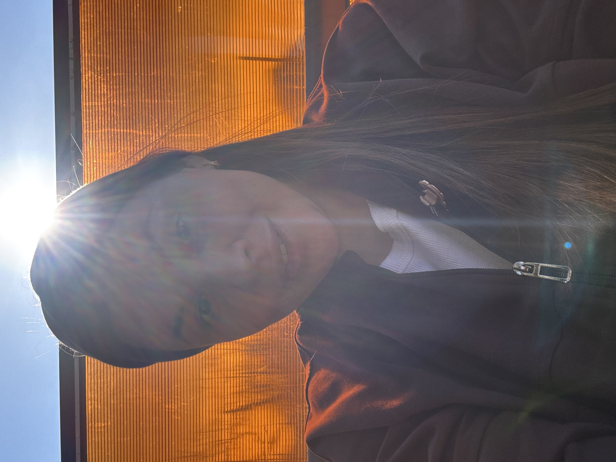 A medium close-up shot depicts a person with long brown hair, wearing a brown zip-up jacket over a white top. The subject's face is partially obscured by bright sunlight. Behind the person is an orange, vertically-lined structure, possibly a wall or fence. Above this structure, a patch of clear blue sky is visible. The scene appears to be outdoors. The location is unidentified.