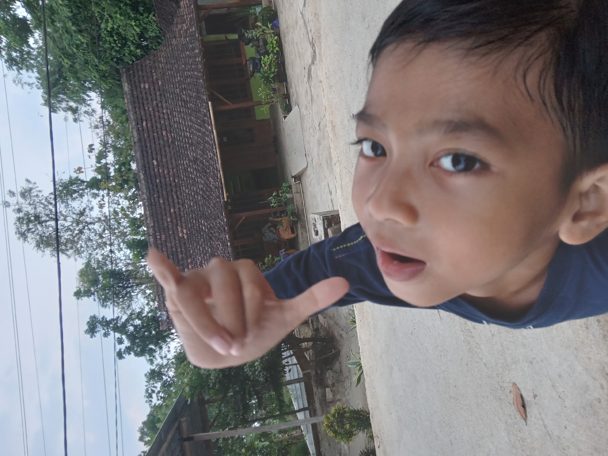 In Kedunggudel, Indonesia, a close-up, Dutch angle shot captures a young person's face and outstretched hand against a backdrop of a rural home and overcast sky. The individual, with dark hair and wide, dark eyes, is wearing a navy blue shirt. Their left hand is raised, with the thumb pointing down, forming a "thumbs down" gesture. The background features a traditional Indonesian house with a tiled roof, wooden beams, and several potted plants. The ground is paved with concrete. The overall scene suggests an outdoor setting during the daytime.