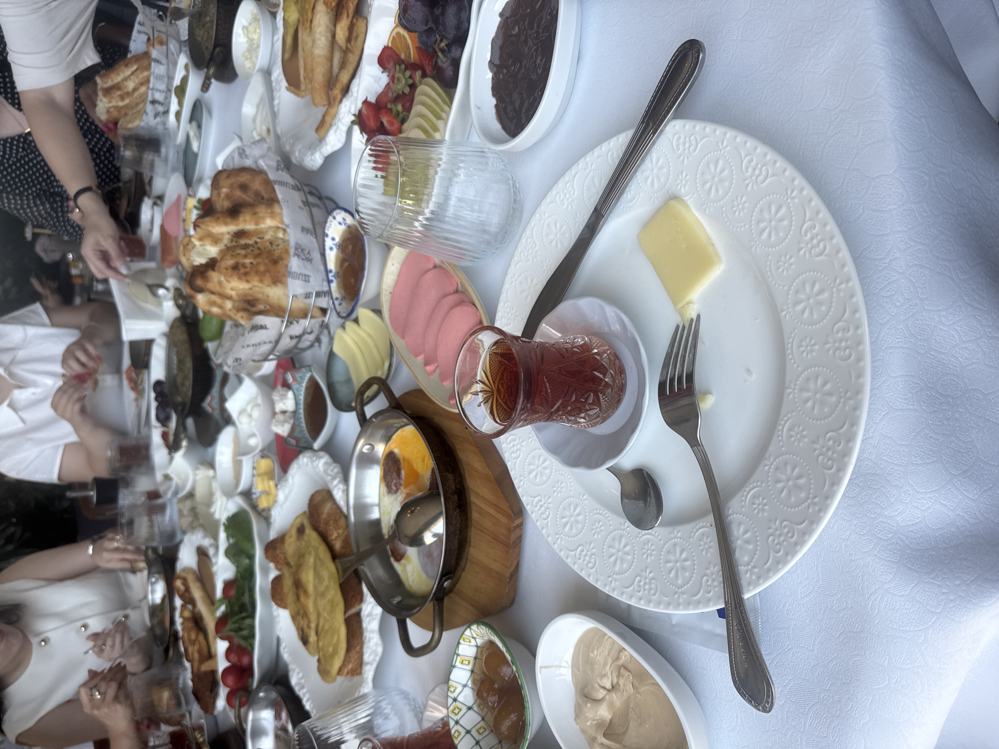 The image depicts a lavish breakfast spread on a white tablecloth, likely in Sabail Raion, Azerbaijan, given the context provided. The table is laden with various dishes, including a pan of fried eggs with sausage, an assortment of breads, cheeses, olives, jams, fruits like strawberries and grapes, and slices of pink bologna. Several glasses of dark tea are also visible, along with cutlery and small bowls containing different spreads and preserves. In the background, blurred figures of people are seated, engaged in eating and conversation. The overall impression is one of a communal, abundant breakfast meal.