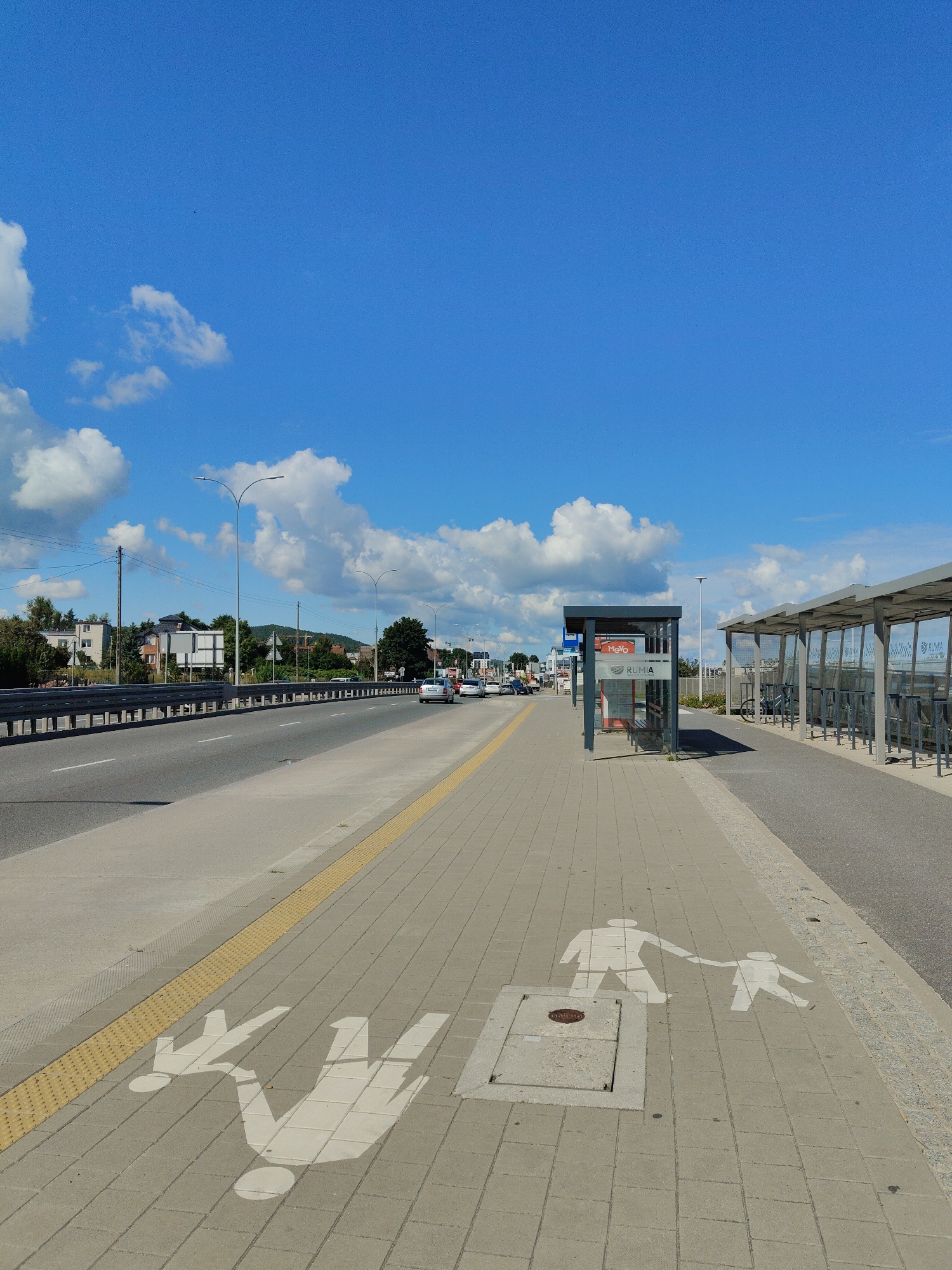 The image depicts a modern bus stop and sidewalk in Rumia, Poland. The bus stop shelters are made of glass and metal, with bicycles parked in the racks beside one of them. The sidewalk is paved with grey bricks and features white stencils of adults holding children's hands, indicating a pedestrian crossing or safety zone. A yellow tactile paving strip runs along the edge of the sidewalk, adjacent to a concrete road with cars driving on it. The sky above is a clear, vibrant blue with scattered white clouds. The overall scene is clean and orderly, suggesting a well-maintained urban environment.