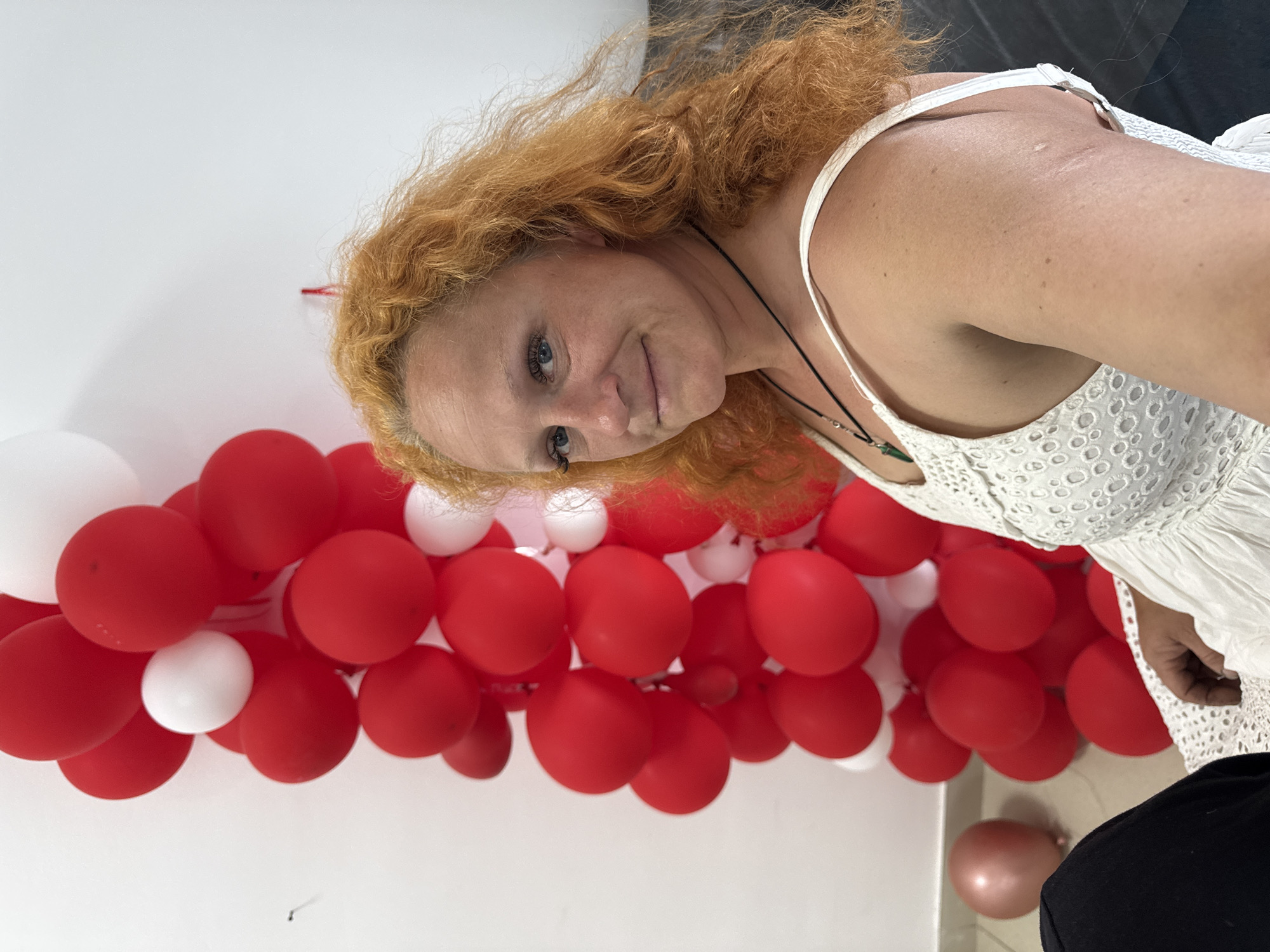 The image shows a woman with vibrant red, curly hair and blue eyes. She is wearing a white, sleeveless dress with intricate lace detailing. She has a necklace with a green pendant. The woman is smiling and looking towards the camera. She is positioned in front of a large column of red and white balloons. To the left of the balloons, a single rose-gold balloon is visible on the floor. The background is a plain white wall, and a dark surface, possibly furniture, is visible to the right. The scene appears to be indoors, possibly at a party or celebration. There is no visual information in the image that can determine the location in Chaikovsky, Russia.