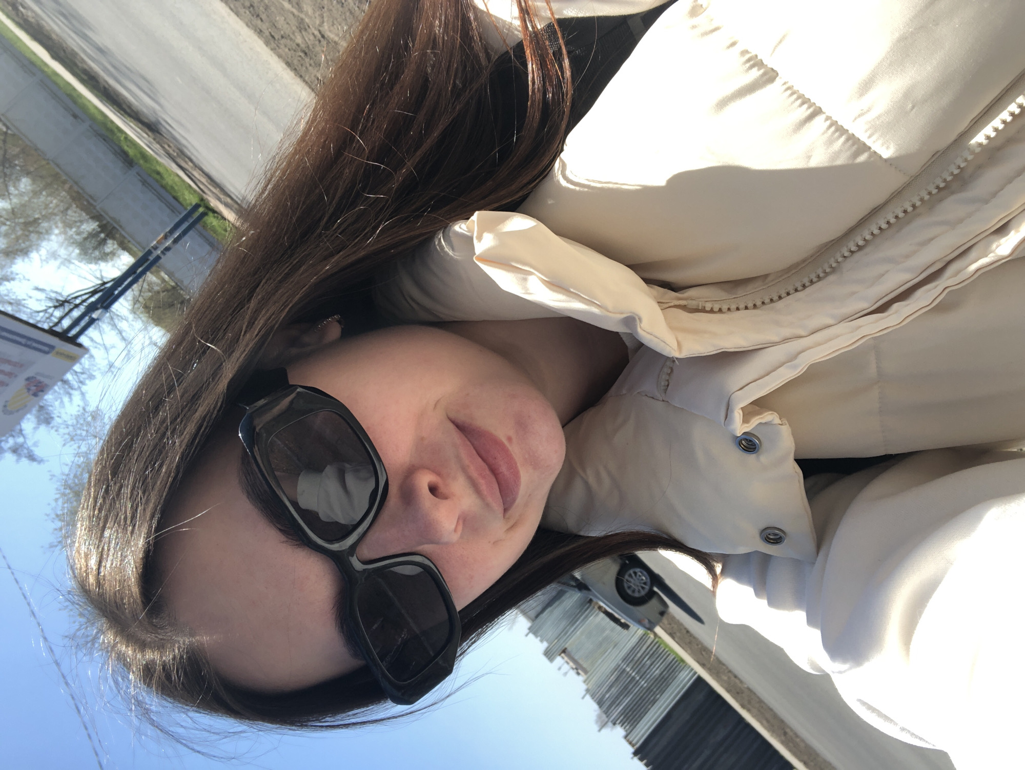 A woman with long, dark brown hair is wearing large, black sunglasses and a light beige puffer jacket. The photo appears to be a selfie, taken outdoors in bright sunlight. In the background, there is a road, trees, a building, and a parked car. Based on the presence of the city-specific street name sign, the location is Chaikovsky, Russia.