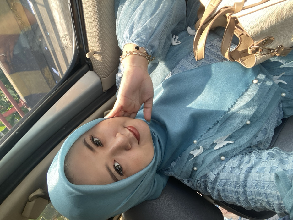 A female subject, appearing to be of Southeast Asian descent, is depicted inside a vehicle in the city of Kebonagung, Indonesia. She is shown from the chest up, looking directly at the camera with a subtle smile. She wears a light teal hijab and a matching dress with sheer, puffed sleeves, adorned with white butterfly appliques and pearls on the bodice. Her right hand is raised, with fingers gently resting against her right cheek. Multiple gold-colored bracelets, one featuring black heart charms, are visible on her right wrist. Her makeup includes defined eyebrows, dark eyeliner, mascara, and reddish-brown lipstick. The interior of the vehicle, including a dark car seat and tan headliner, is visible. Through the vehicle's right window, bright daylight illuminates a partial view of an outdoor environment, revealing a red railing and green foliage. A beige handbag with brown accents rests on the right side of the frame, near the subject.