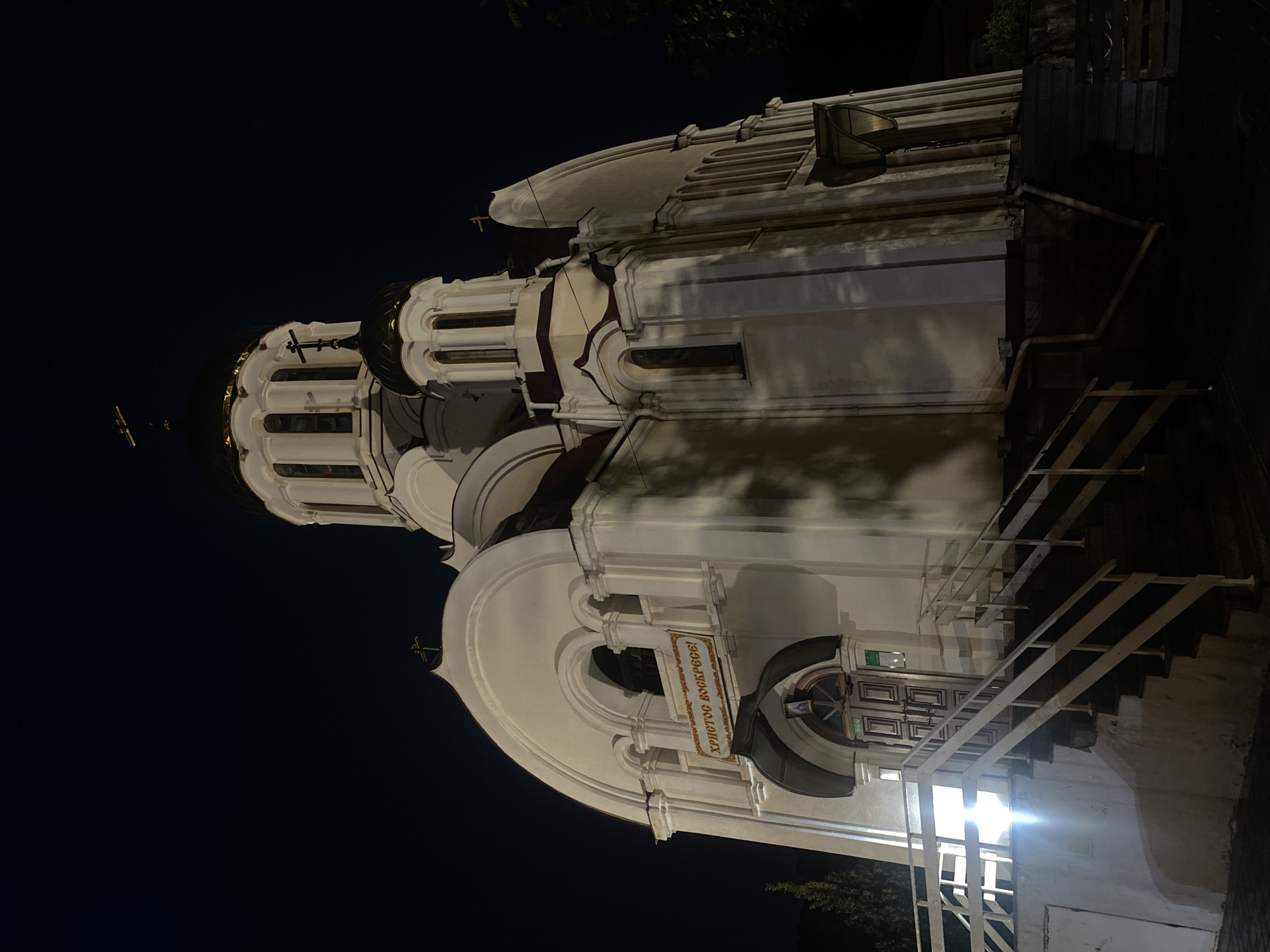 This is a vertical, nighttime, outdoor photograph of an Orthodox church. The church is illuminated by artificial lights, casting shadows and highlighting its white facade. The architecture features multiple domes, arched windows, and decorative cornices. A golden cross is visible atop one of the domes. A sign with Cyrillic text, "ХРИСТОС ВОСКРЕСЕ!" (Christ is Risen!), is prominently displayed near the entrance. The entrance itself has a large, arched doorway with wooden double doors. A white metal staircase leads up to the entrance. The surrounding area is dark, with only the church and its immediate vicinity illuminated, suggesting a clear night. The image is taken from a low angle, looking up at the church's imposing structure. The scene evokes a sense of solemnity and religious reverence.