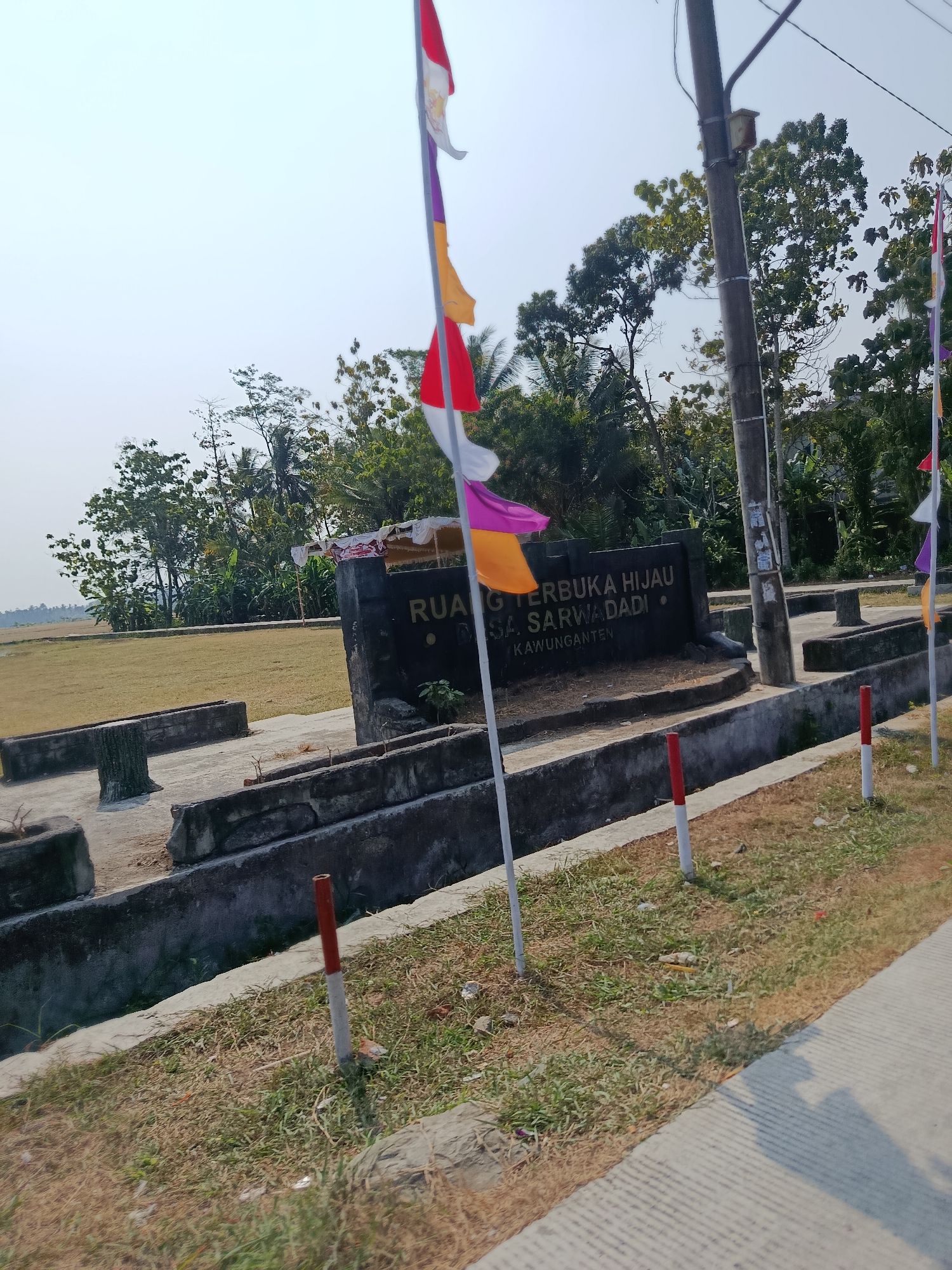 The image shows a monument with text that reads "RUANG TERBUKA HIJAU SARWADADI". The monument is located in Sarwadadi, Indonesia, and is surrounded by a grassy area and some concrete structures. Several flags, including Indonesian flags, are visible in the foreground. The sky is clear and the sun is shining, casting shadows on the ground. Trees and vegetation are present in the background.