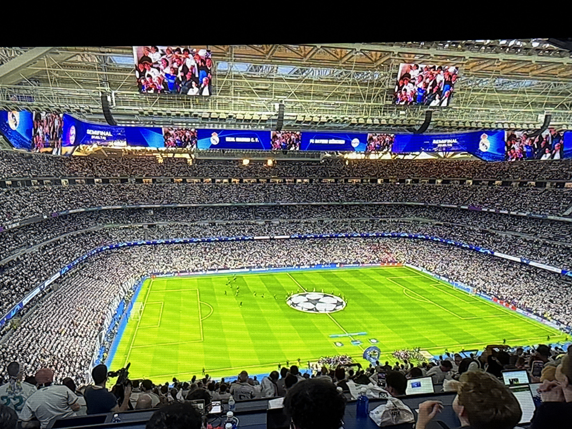 The image shows a large crowd in a stadium, most likely for a soccer game. The field is green with a Champions League logo in the center. There are screens displaying the game and team logos. The audience is densely packed in the stands. People are mostly sitting and watching the game. The stadium has a closed roof. The location is not in Sofia, Bulgaria.