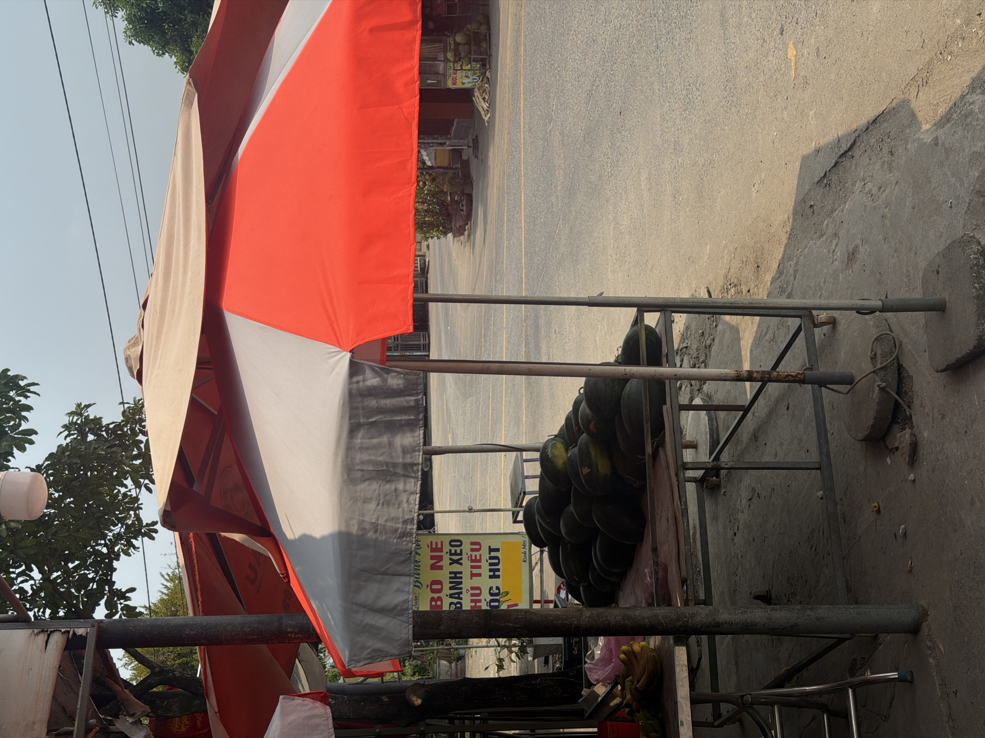 The image shows a street vendor in Đà Nẵng, Vietnam. A large stack of watermelons sits on a metal cart under an orange and grey striped umbrella. A sign in Vietnamese is visible, indicating the vendor sells "Bò Né," a sizzling beef dish, along with other items. The cart is situated on a concrete surface next to a paved road. A curb separates the road from the vendor's location. The setting appears to be an open-air environment. Trees are visible in the background and to the left. Power lines run above.