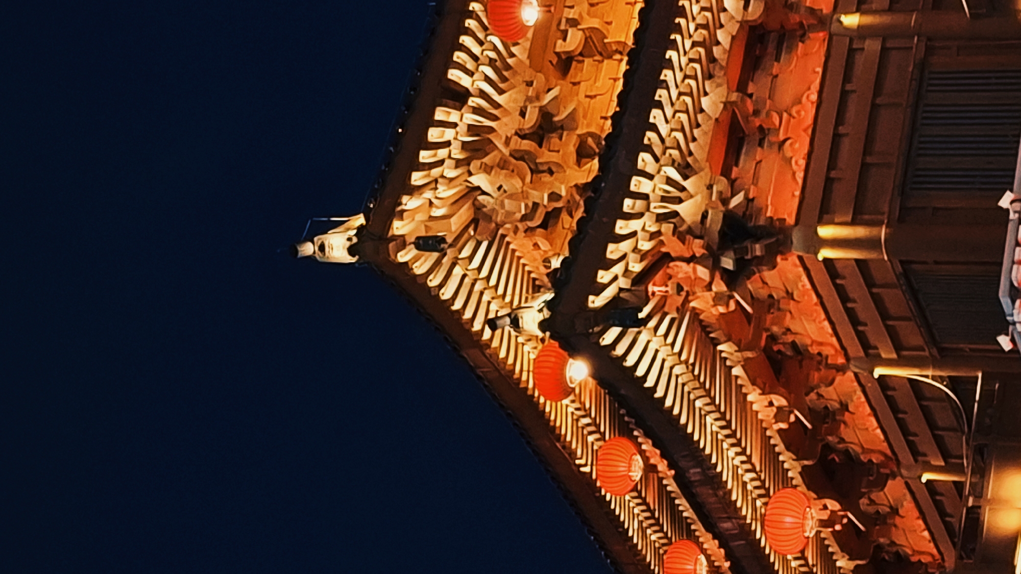 This close-up shot captures the roof of a building in Yanta District, China. The architecture is ornate, featuring stacked, layered elements. The roof is brightly illuminated with warm lights, with strings of red lanterns hanging from the eaves. The sky is a dark, clear navy blue.