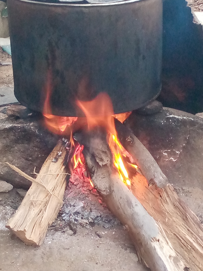 A large, dark metal pot is suspended over a fire pit in Ajaokuta, Nigeria. The fire is burning with bright orange and yellow flames, illuminating the underside of the pot and casting a warm glow on the surrounding area. Several pieces of firewood are visible, some charred and smoldering. The ground is covered in sand and ash. The scene appears to be outdoors, with a rustic and simple setup for cooking.
