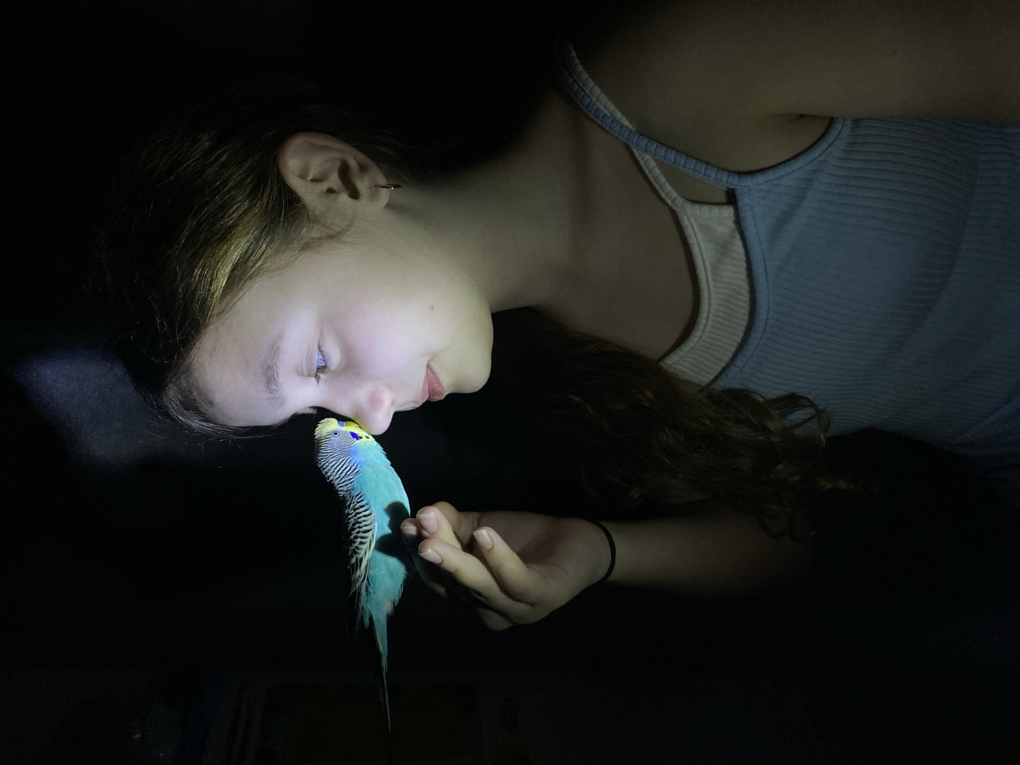 A young female lies in a darkened environment, possibly a bed or couch given the soft appearance of the unseen surface. She wears a light blue ribbed tank top with white straps. Her long brown hair is spread beside her. She is angled downward with her face close to a blue and yellow budgerigar (parakeet) perched on her hand. A direct, bright light source is present, illuminating her face, the bird, and her hand. The rest of the environment is largely obscured by darkness.