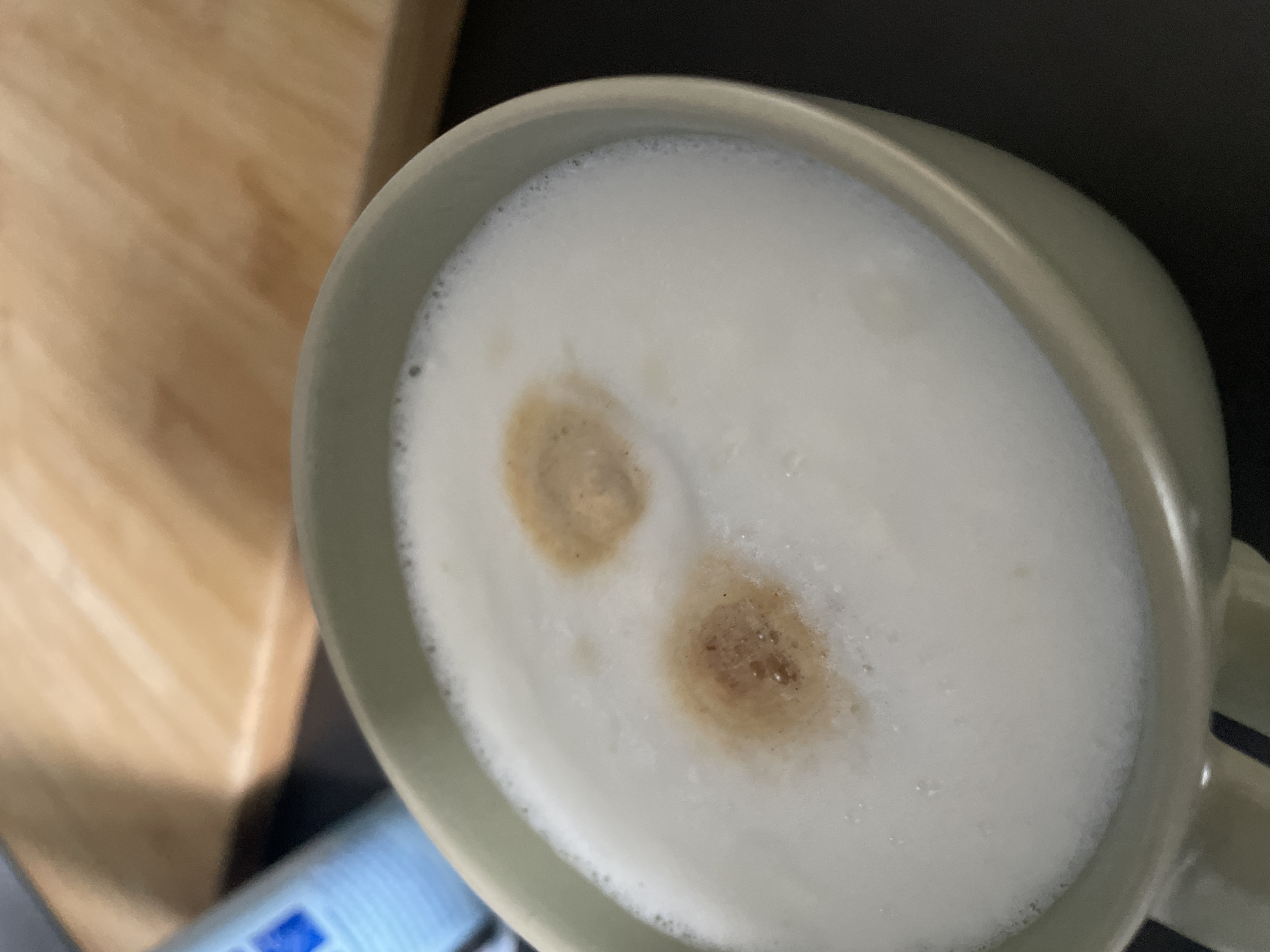 The image shows a close-up of a cup of coffee with a foamy top and two brown spots of espresso. The cup is light green and appears to be made of ceramic. The coffee is in a setting that suggests a domestic or cafe environment, with a wooden surface in the background and a dark countertop below. There are no people visible in the image. The scene is lit with natural light, casting soft shadows. The image does not provide any information about the city of Morsbach, Germany.