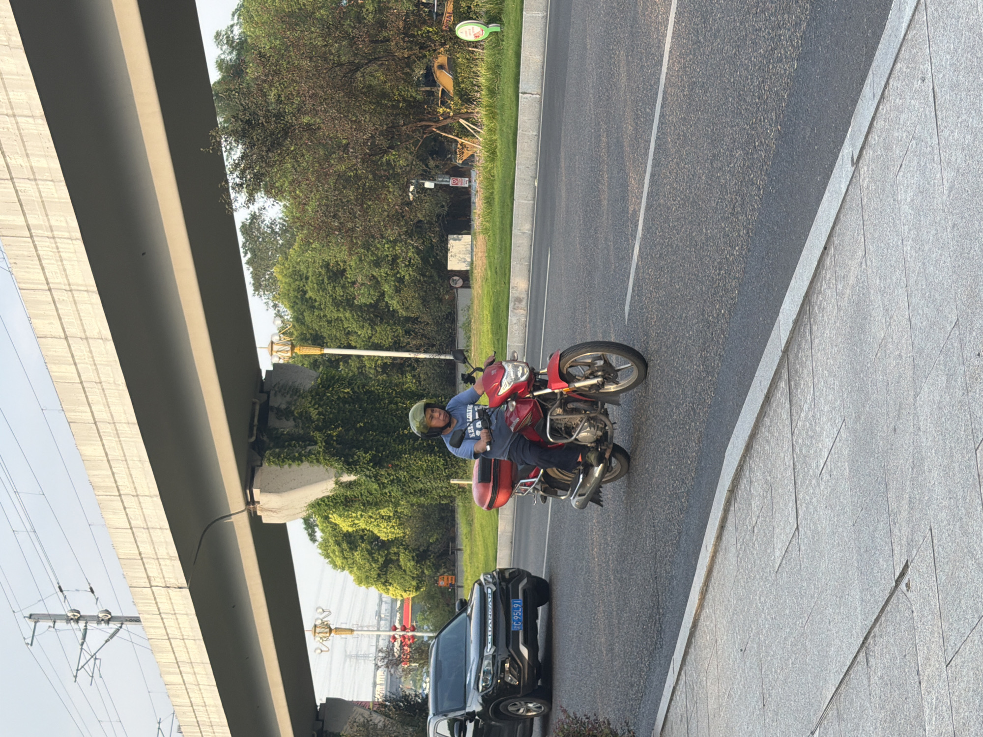 A man wearing a helmet and a blue t-shirt rides a red motorcycle with a red storage box on the back along a paved road. He is in motion, and the motorcycle is angled towards the right of the frame. To the left of the motorcycle, a black SUV with a license plate reading "C 95L91" is also on the road. Both vehicles appear to be traveling in the same direction. In the background, a large concrete overpass with visible structural supports and overhead electrical wires looms above. Lush green trees and vegetation line the side of the road, and in the far background, industrial structures and possibly construction equipment are visible. The scene appears to be in an urban or suburban area with elevated infrastructure, consistent with a location in the city of Jiangjin, China. The lighting suggests it is daytime, possibly late afternoon.