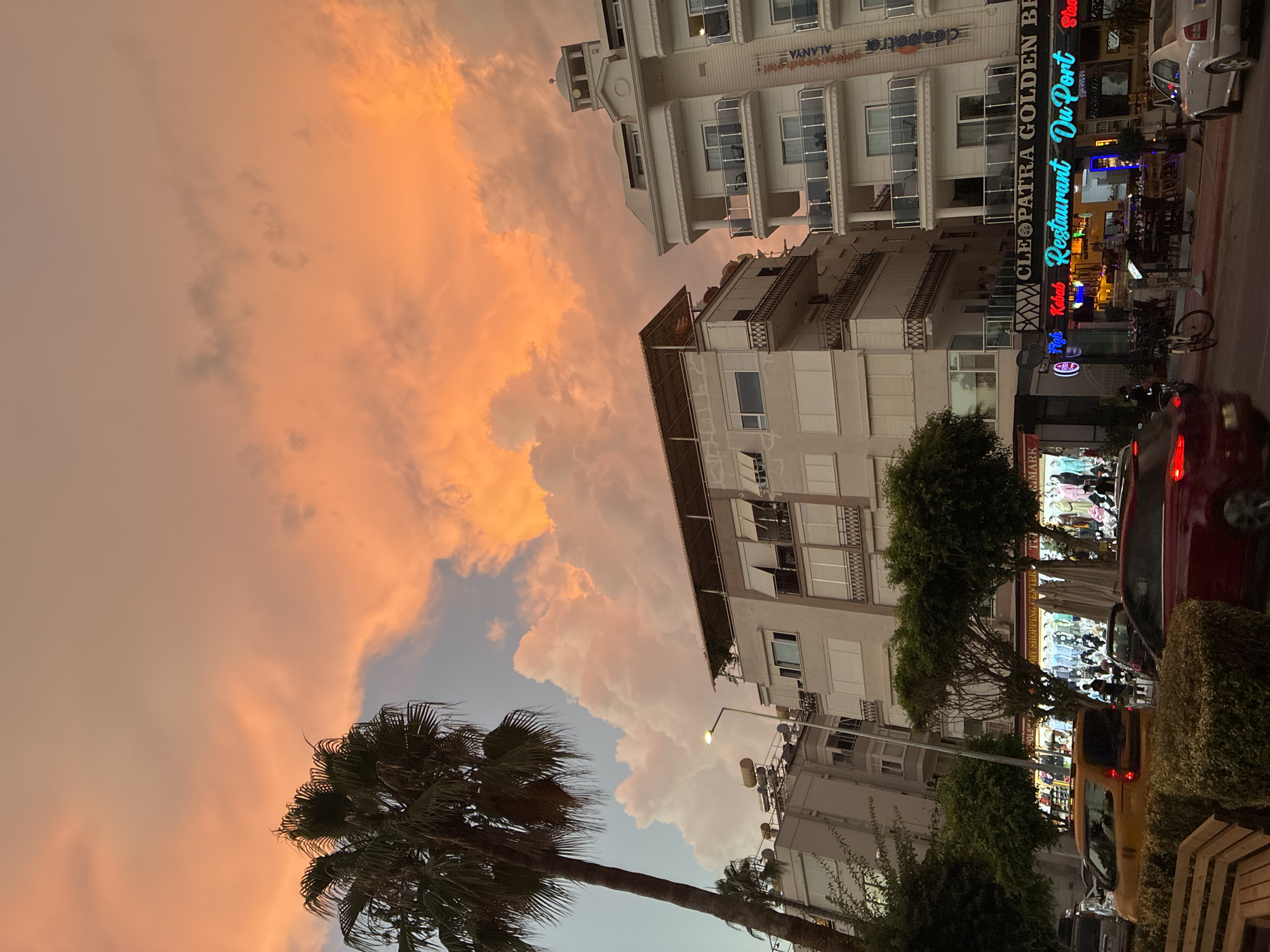 The image depicts a street scene in Alanya, Turkey, at dusk. The sky is filled with dramatic orange and pink clouds, suggesting a sunset. In the foreground, a busy street is lined with buildings, shops, and restaurants. A palm tree stands prominently on the left side of the image.
Several buildings are visible, including apartments and what appears to be a hotel or resort on the right, with balconies and white exteriors. Below these buildings are shops and restaurants. One establishment is identified as "CLEOPATRA GOLDEN BEER RESTAURANT DU PORT".
Various vehicles are present on the street, including a yellow taxi, a red car, and a silver car. Pedestrians can be seen walking on the sidewalks and near the shops. A bicycle is parked on the side of the street. The shops display various goods, and the restaurants have outdoor seating areas. The overall atmosphere is that of a lively urban environment transitioning into the evening.
