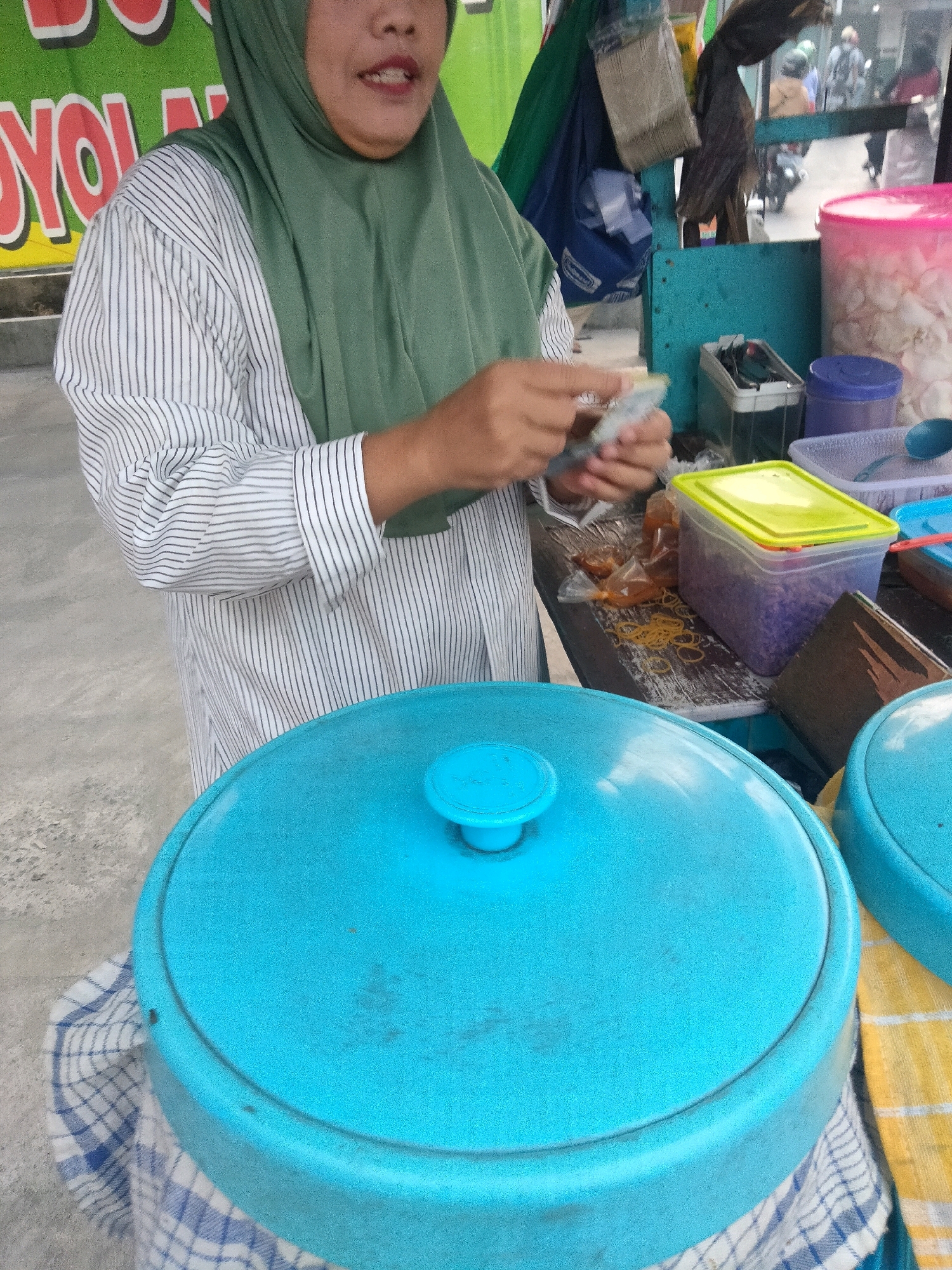 In Pisangan Jaya, Indonesia, a woman wearing a green hijab and a striped shirt is captured in a medium shot at what appears to be a street food stall. She is standing behind a counter, her hands in motion as she prepares food, holding a small plastic bag with some contents. The background reveals a vibrant green banner with red text, partially obscured. To her right, a blue stall structure is visible, with various containers of food items, including a large pink container filled with white food. Below the counter, two large blue containers with lids are prominent. One is covered with a blue and white checkered cloth, and the other is partially visible to the right, also covered with a cloth, this one yellow and white checkered. The scene suggests a busy moment at a local food vendor.