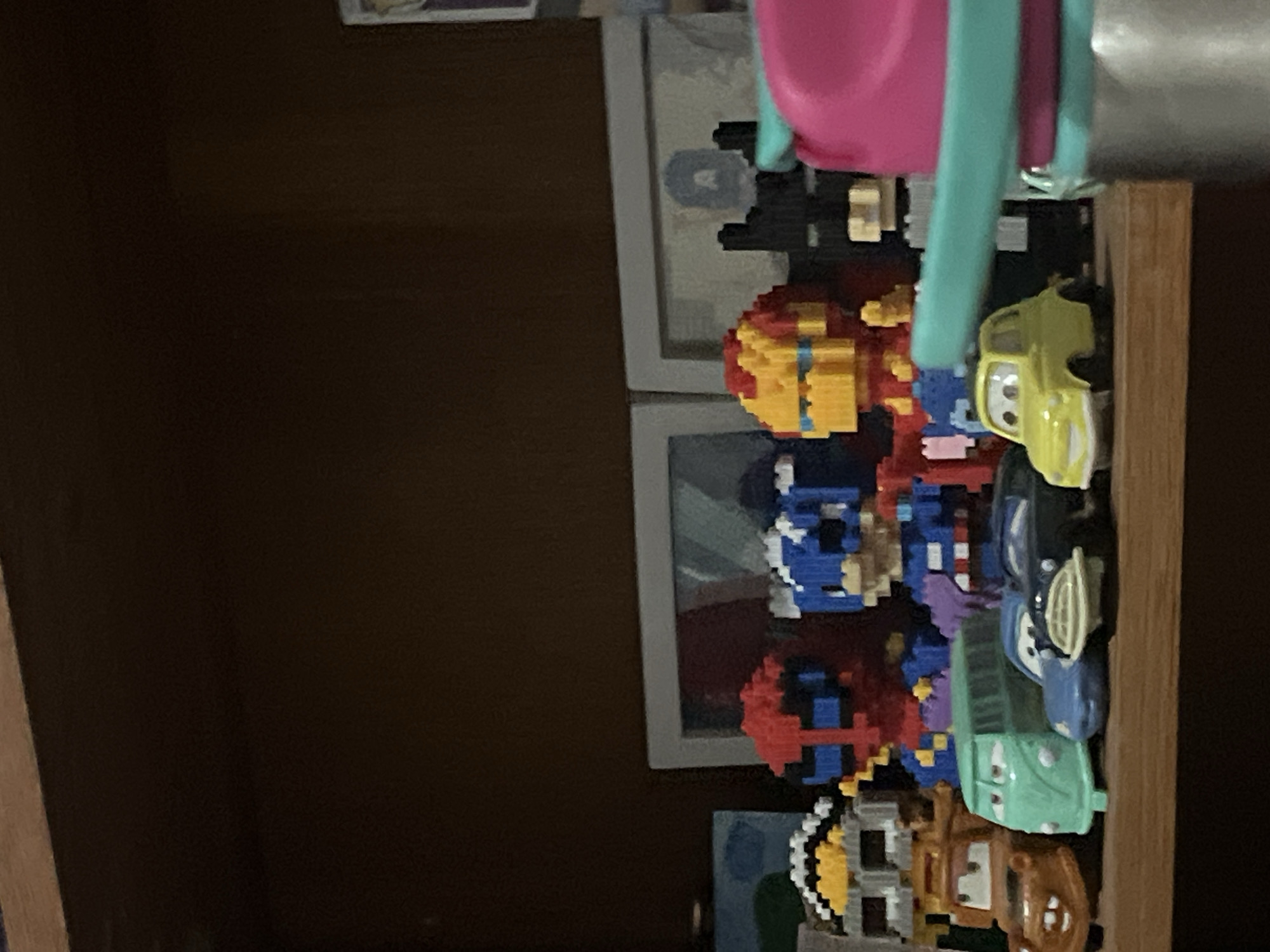 This image depicts a shelf with various collectible toys. On the shelf, there are several figures that appear to be constructed from small building blocks, resembling characters from popular superhero franchises like Spider-Man, Captain America, and Iron Man. In front of these figures, there are miniature toy cars, including a brown tow truck, a light green van, a dark blue car, and a small yellow car. The background shows what appears to be framed pictures on the wall behind the shelf. The lighting suggests an indoor setting, likely a home or a display area. The shelf itself is made of wood. The image does not provide any specific information or visual cues that would allow for the identification of its location in Santa Ana, Venezuela.