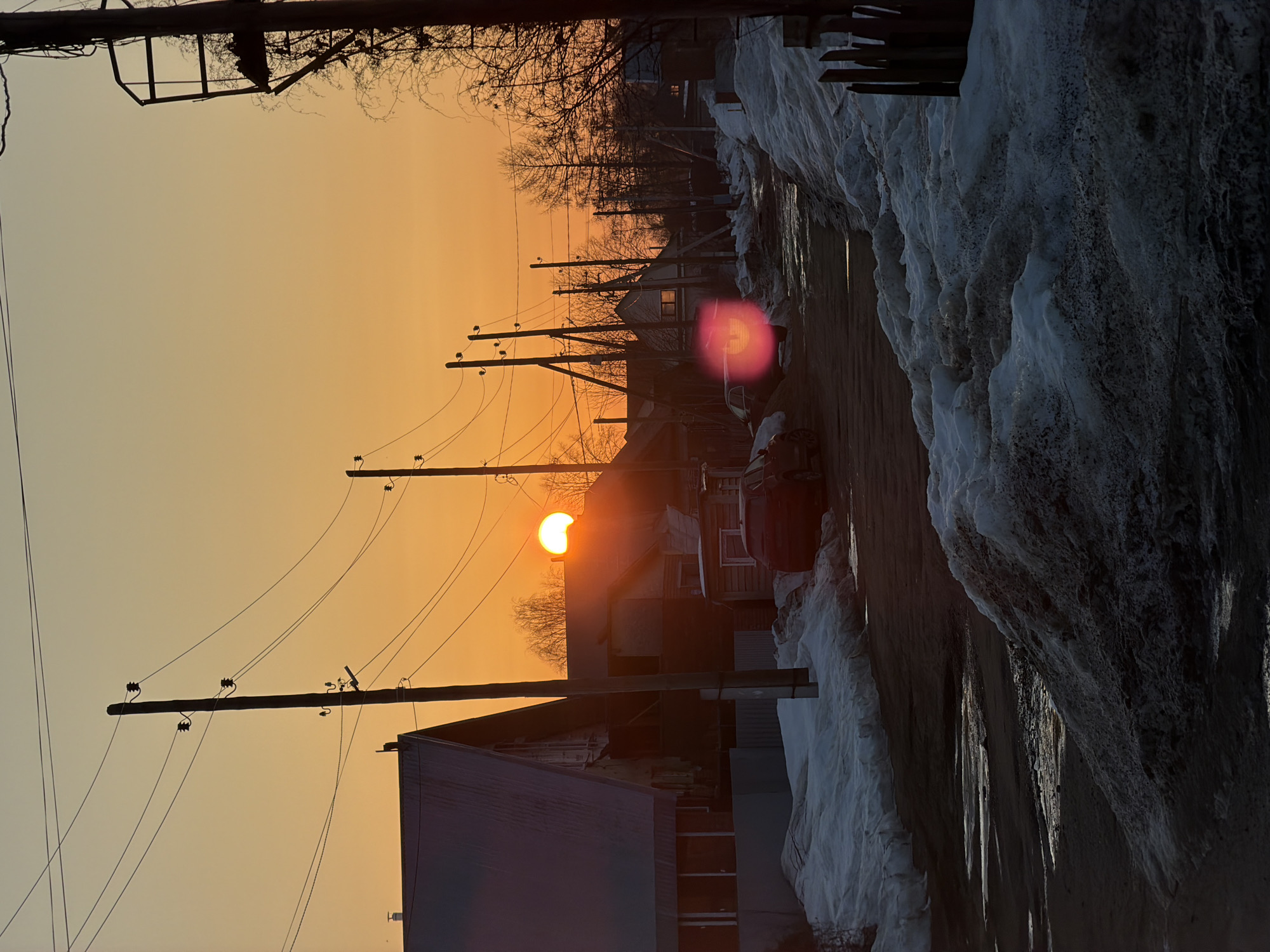 The image depicts a residential street scene in Chaikovsky, Russia, at either sunrise or sunset, evidenced by the low, orange sun partially visible above the horizon on the left and the warm gradient of the sky. The environment is characterized by persistent snow cover, with large, dirty snowbanks lining both sides of the unpaved or damaged road surface, which shows signs of melting with reflective puddles.
The foreground features a significant accumulation of dark, melting snow on the right, alongside what appears to be part of a wooden fence. Several wooden utility poles with multiple overhead power lines are prominently silhouetted against the bright sky, extending down the street. On the left, several detached houses are visible; the closest is a large structure with a prominent gable roof, partially obscured by a utility pole. Further down the street, other residential buildings, primarily of single or two-story construction, are discernable. Bare deciduous trees and some evergreens are visible in the background, between and behind the houses.
A dark-colored sport utility vehicle (SUV) is parked on the left side of the street, partially obscured by a snowbank and another utility pole. A distinct pink-red lens flare is present in the middle ground, near where another vehicle's rear lights might be. No individuals are visible within the frame. The overall impression is a transitional season, likely late winter or early spring, characterized by melting snow and clear, low-angle sunlight.