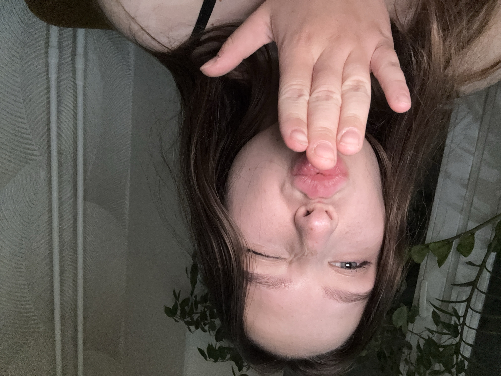 A young woman, likely in her late teens or early twenties, is depicted in a close-up, chest-up shot. She has fair skin with visible freckles on her nose and cheeks, and long, dark brown hair that frames her face. Her left eye is open, revealing a blue-grey iris, while her right eye is closed in a wink. Her lips are pursed into a kissing shape. Her right hand is positioned in front of her mouth, palm outward, with fingers slightly curled, in the action of blowing a kiss. A dark strap of clothing is visible on her right shoulder. To the left of the woman, a dark window frame is partially visible, obscured by the green leaves of what appears to be an indoor plant. To the right, the background consists of a light-colored wall or curtain featuring a subtle textured pattern of faint vertical lines and a wavy or herringbone motif. The overall lighting is dim, with the woman's face adequately illuminated. The depicted scene is located in what is stated to be the city of Chaikovsky, Russia.