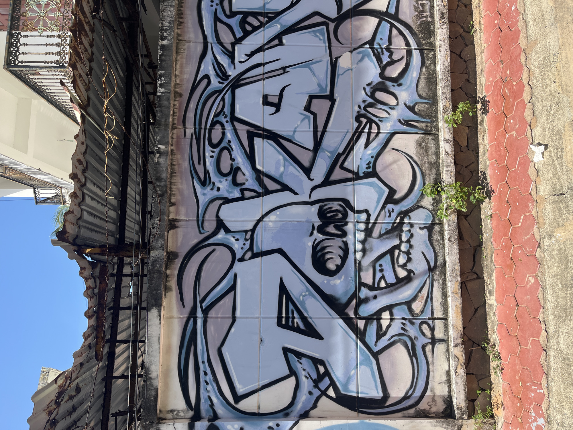A close-up, eye-level shot captures graffiti on a concrete wall in Mũi Né, Vietnam. The graffiti, rendered in shades of light blue and black, features abstract, stylized lettering that appears to spell out "DAZA." A skull-like motif is integrated into the artwork. Above the wall, a rusted, corrugated metal roof is supported by dark beams and partially obscured by barbed wire. A balcony with decorative iron railings is visible on a building in the background. The ground in the foreground is paved with red hexagonal tiles, transitioning to a concrete sidewalk. Small green plants sprout from the base of the wall and along the edge of the tiled area. The sky is a clear, bright blue.