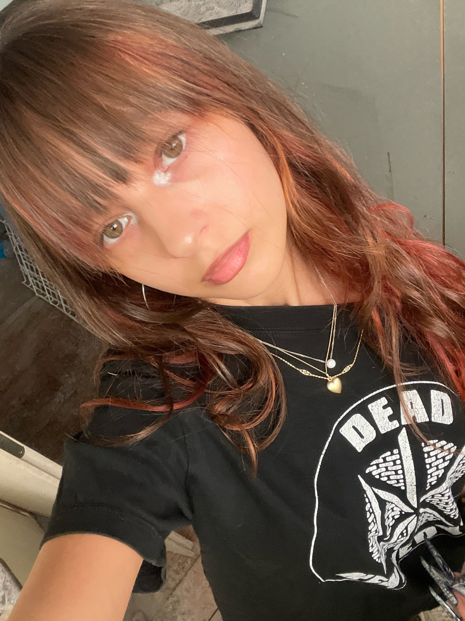 A young person with long, wavy brown hair with red highlights and bangs, wearing a black t-shirt with a white graphic that reads "DEAD" and depicts a star-like design, is shown in a close-up, angled selfie. The person has light skin, brown eyes, and is wearing two delicate gold necklaces, one with a small heart pendant. The background is out of focus, appearing to be an indoor setting with muted colors and textures that could suggest a home or studio environment. The lighting is soft, creating gentle highlights on the face and hair.